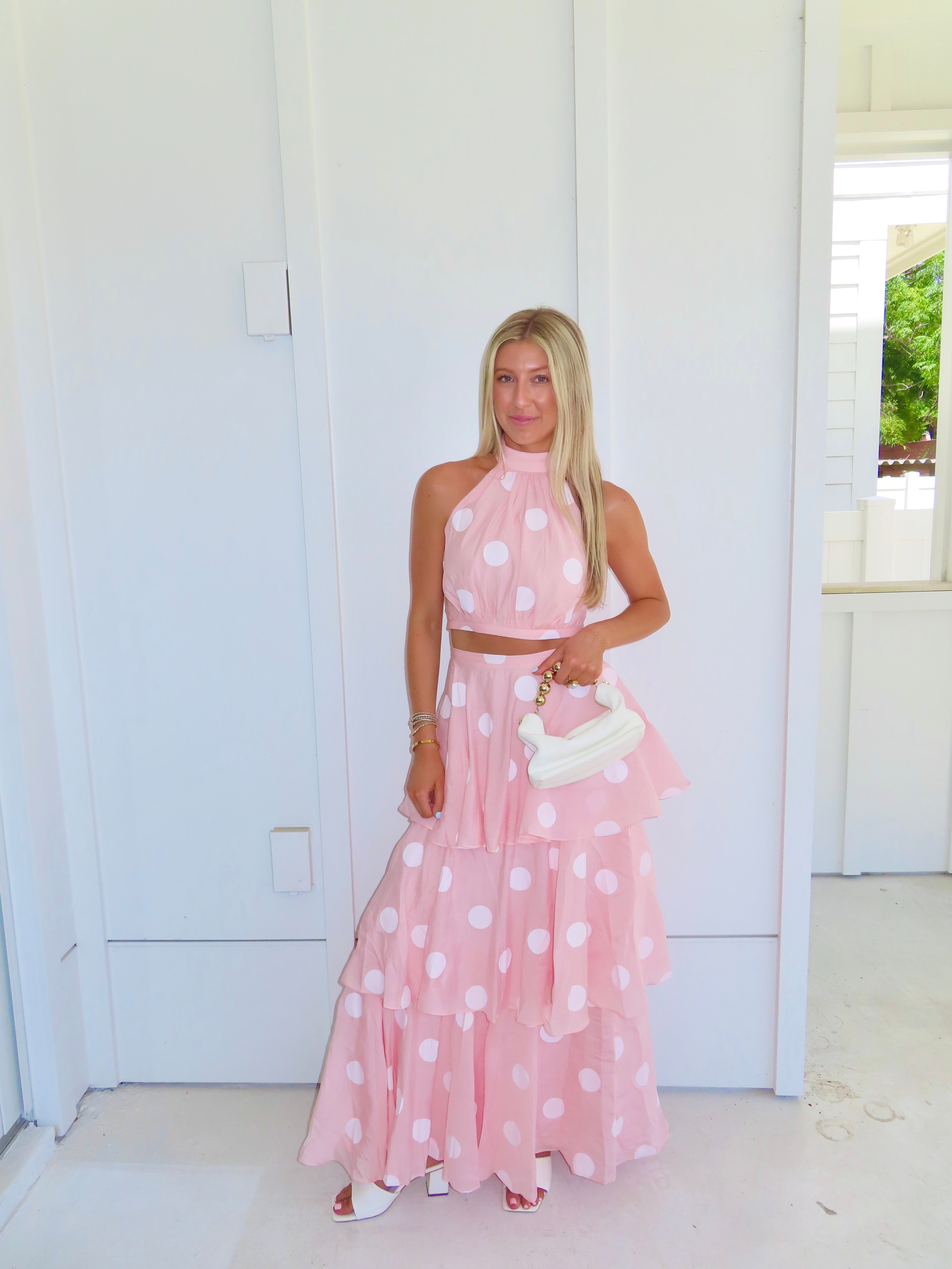 Woman wearing a pink polka dot skirt set holding a white clutch on a bright white patio.