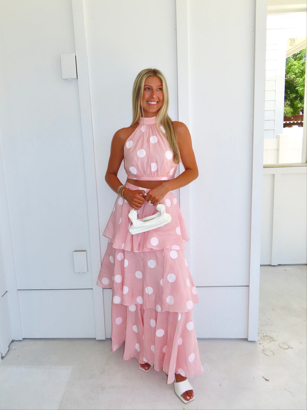 Woman wearing a pink polka dot skirt set holding a white clutch on a bright white patio.