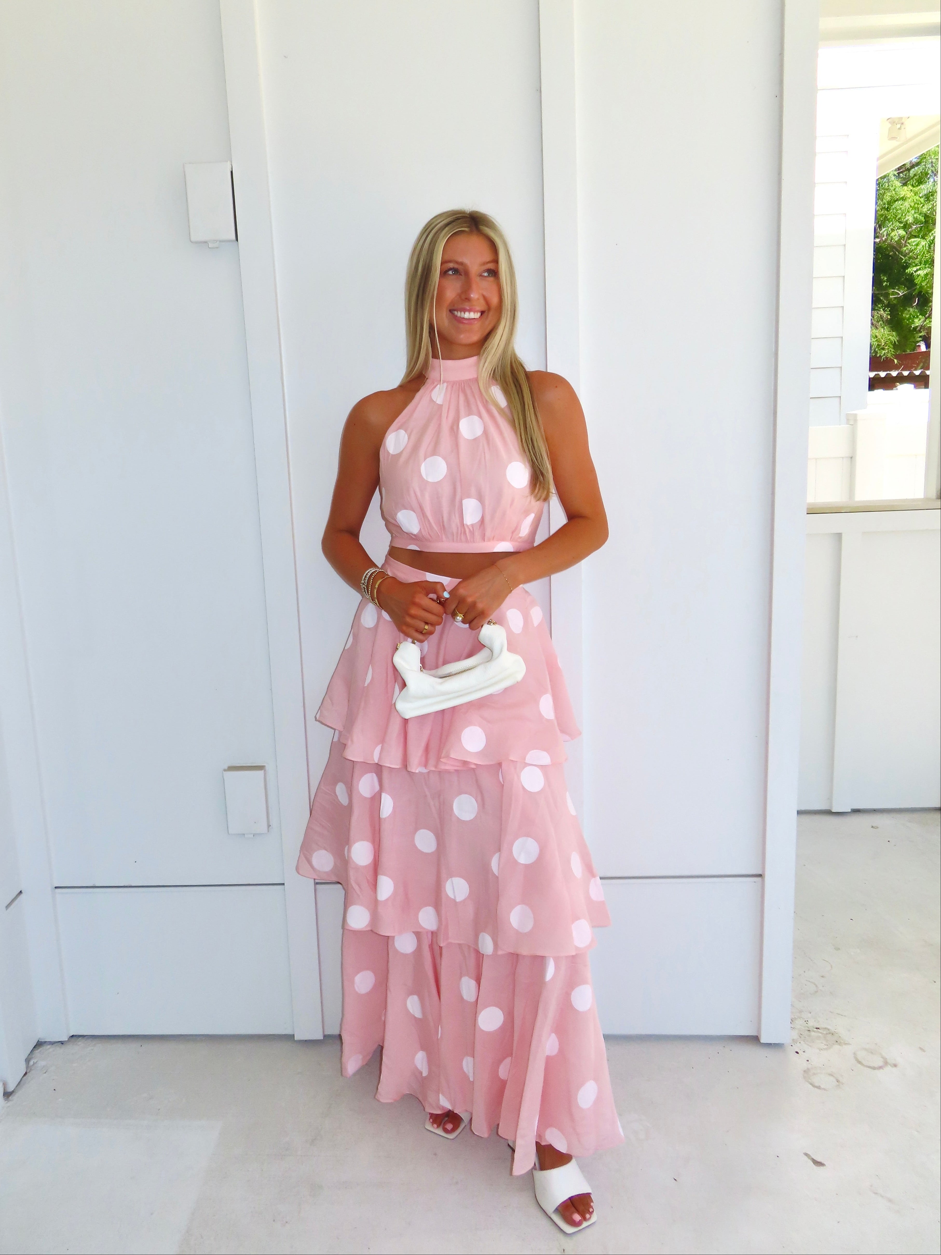 Woman wearing a pink polka dot skirt set holding a white clutch on a bright white patio.