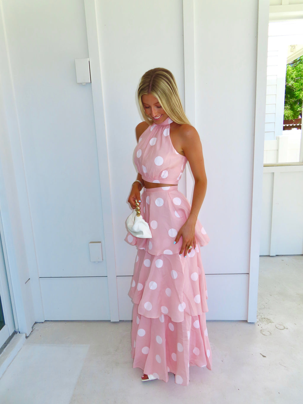Woman wearing a pink polka dot skirt set holding a white clutch on a bright white patio.