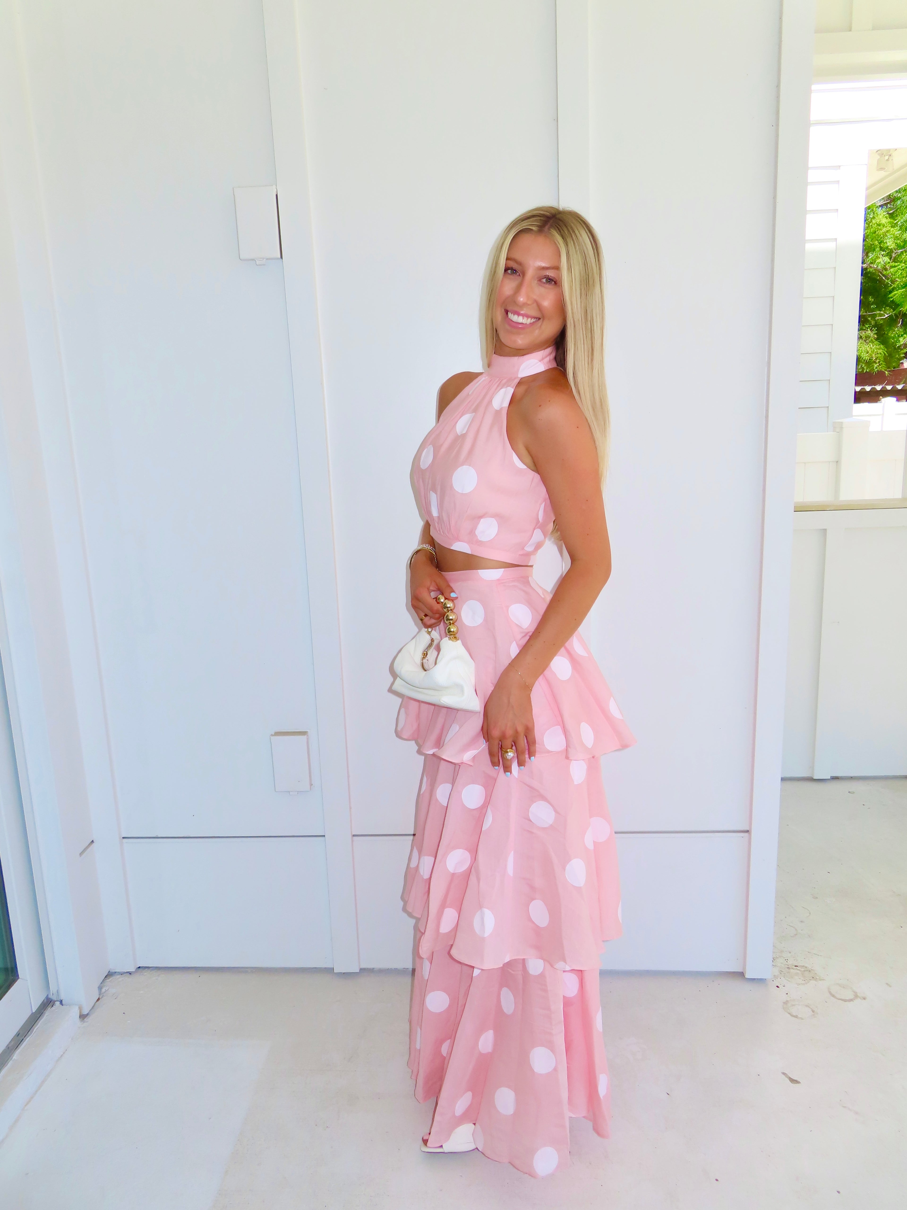 Woman wearing a pink polka dot skirt set holding a white clutch on a bright white patio.