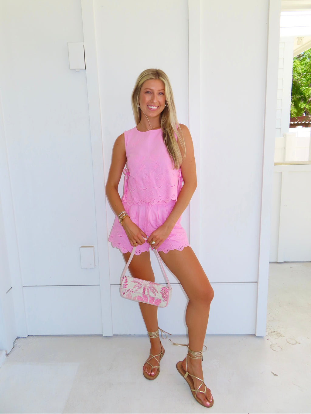 Woman in a pink eyelet shorts set holding a pink and white purse standing on a patio with white walls.