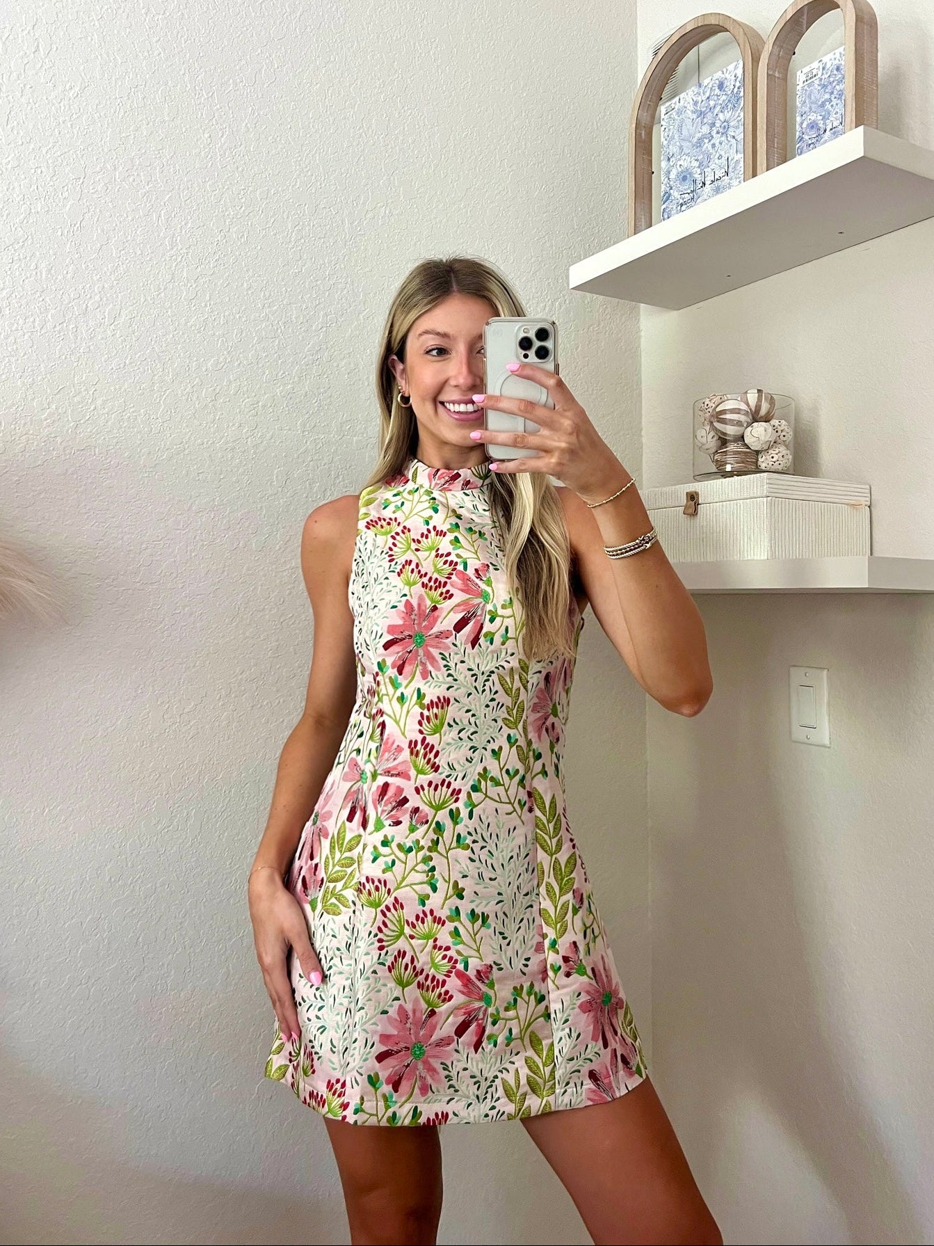 Woman taking a mirror selfie wearing a floral high neck mini dress in a minimalistic room.
