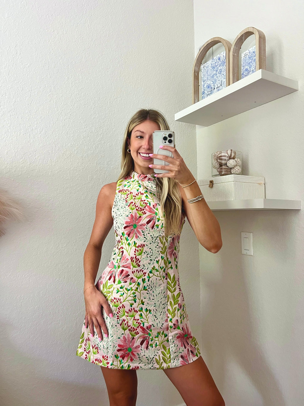 Woman taking a mirror selfie wearing a floral high neck mini dress in a minimalistic room.