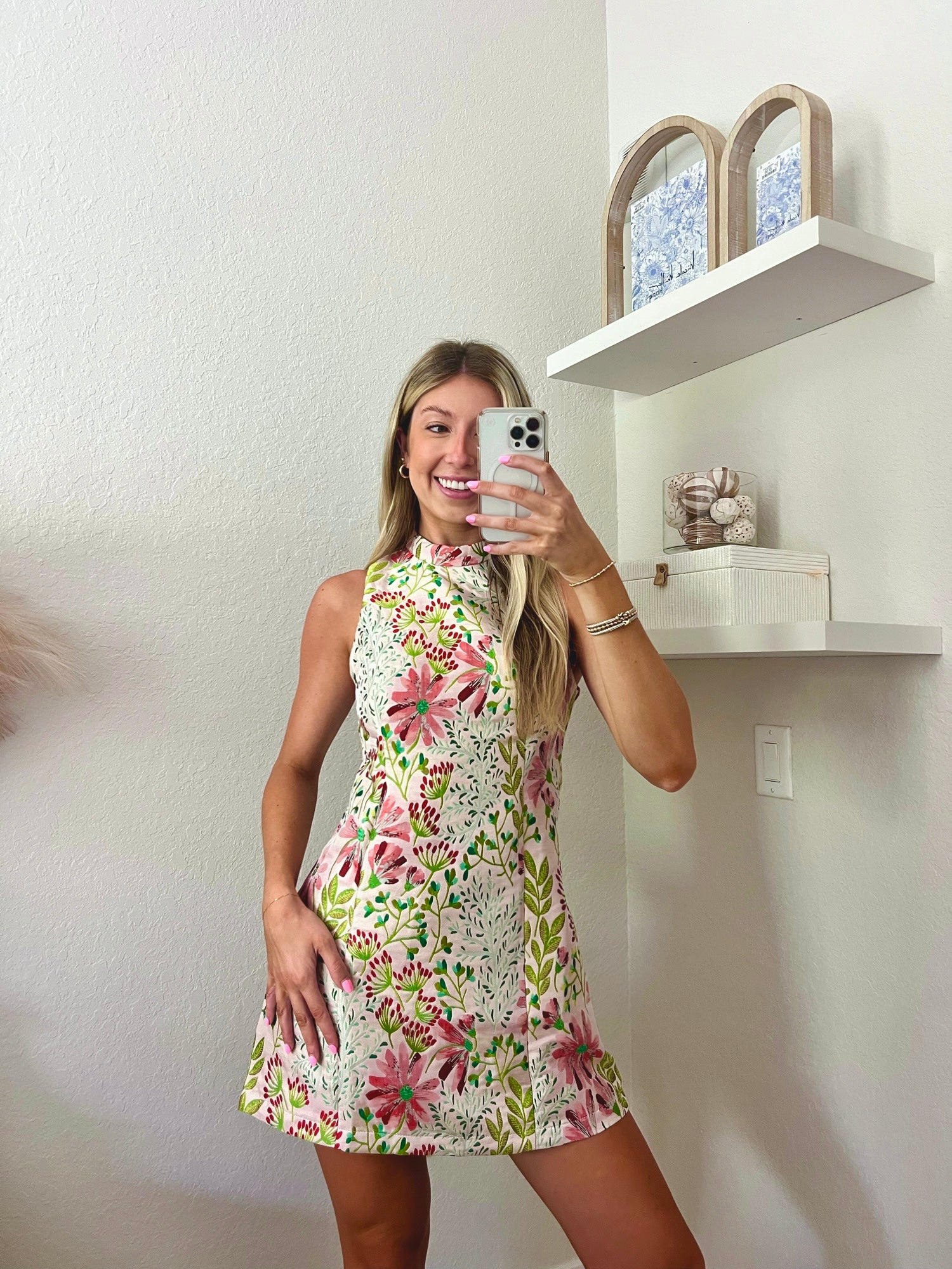 Woman taking a mirror selfie wearing a floral high neck mini dress in a minimalistic room.