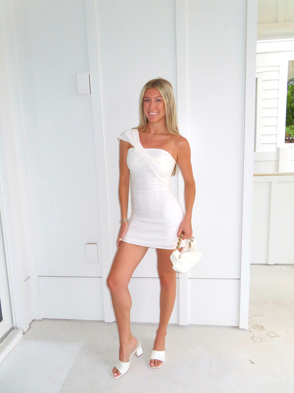 Woman in a white one-shoulder dress holding a clutch and wearing heels against a white wall.