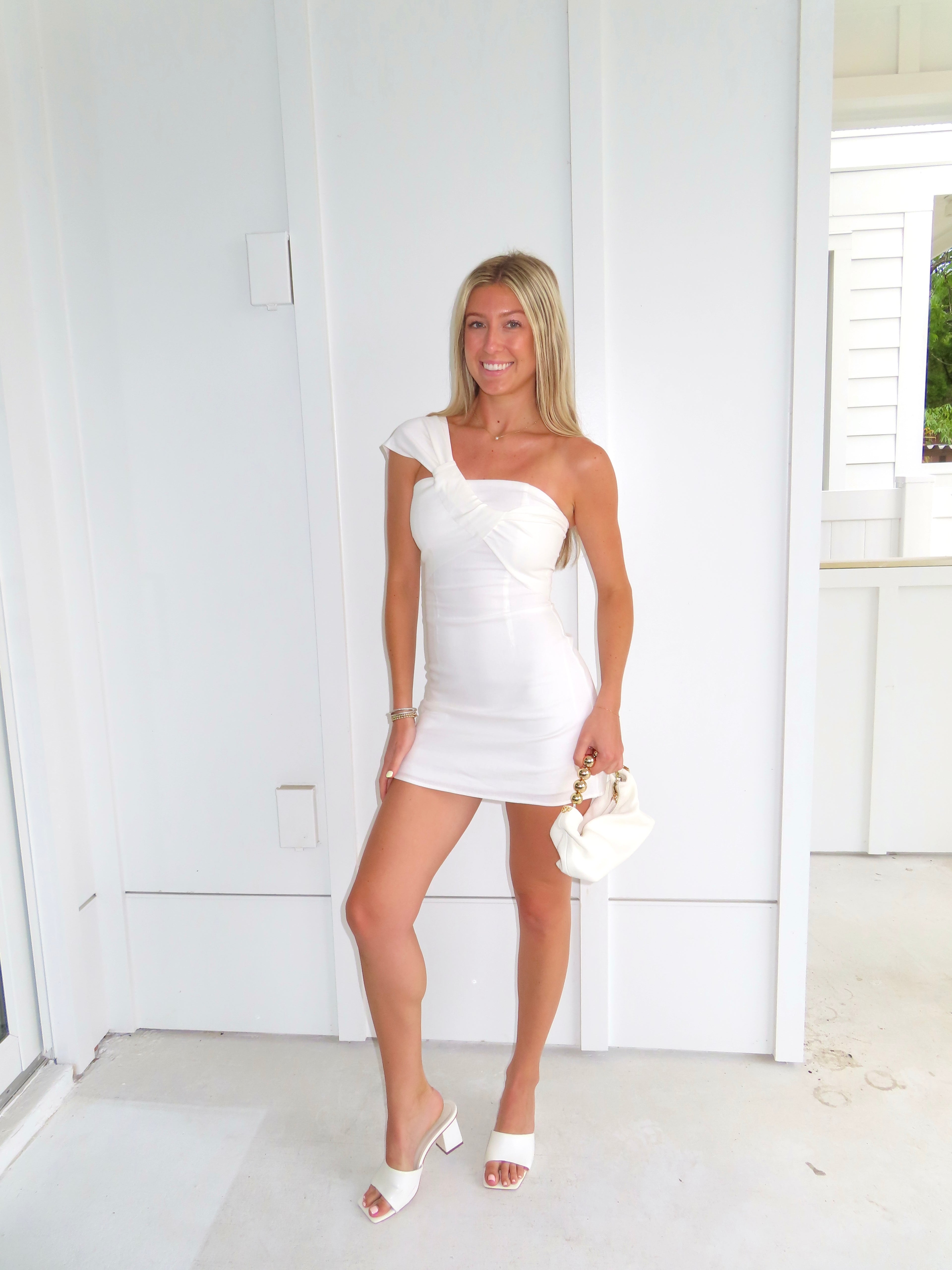 Woman in a white one-shoulder dress holding a clutch and wearing heels against a white wall.
