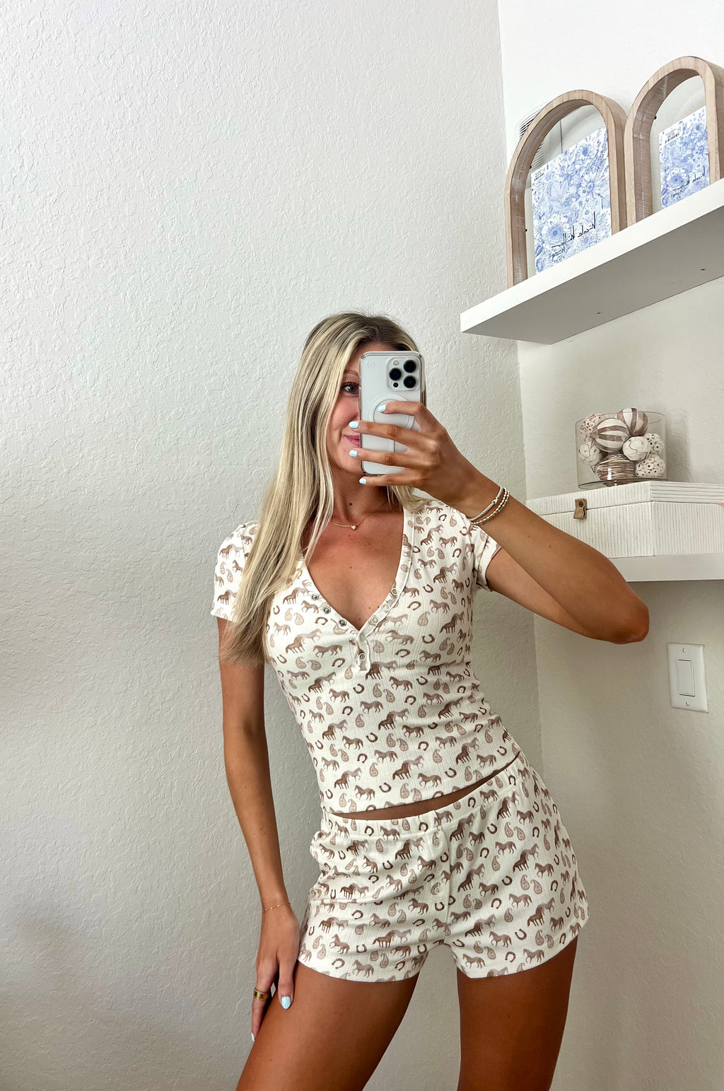 Woman taking a mirror selfie wearing an ivory lounge set with western graphics on it in a room.