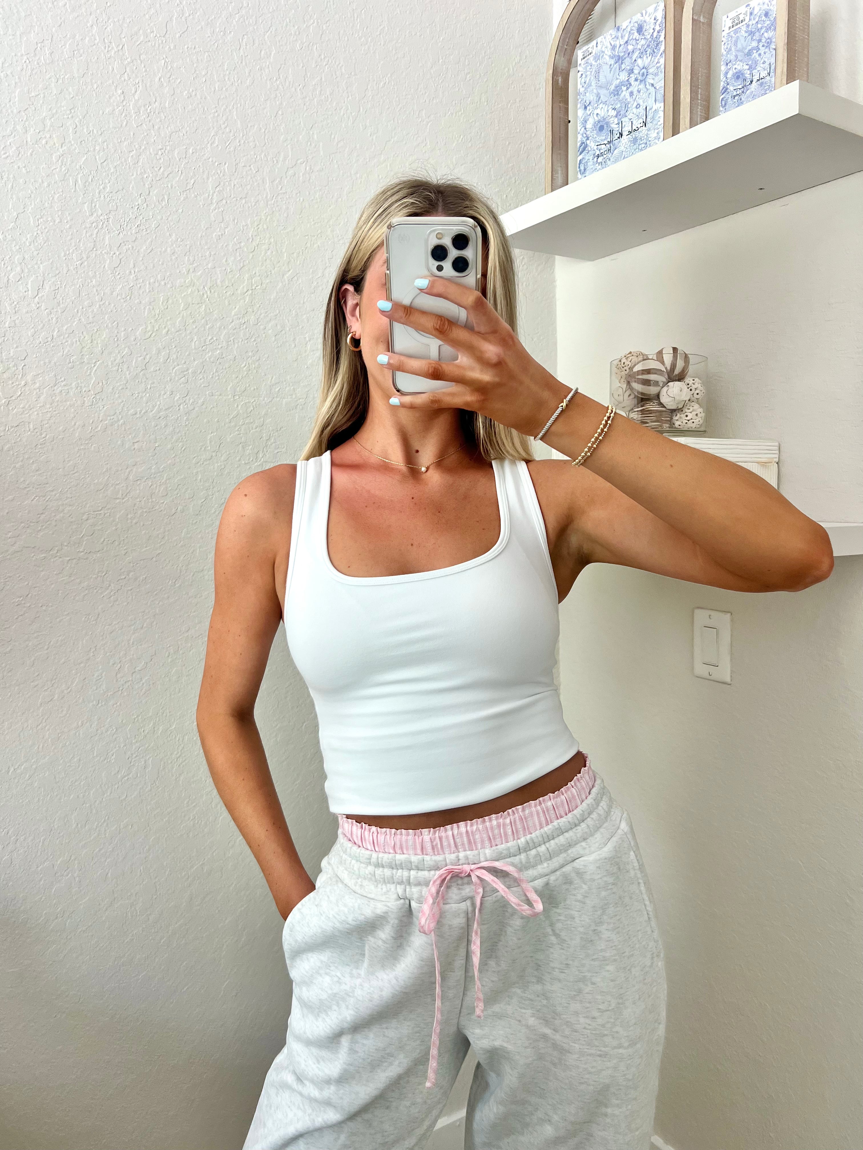 Woman taking a mirror selfie wearing a white tank top and light gray sweatpants with pink and white boxers underneath in a room.