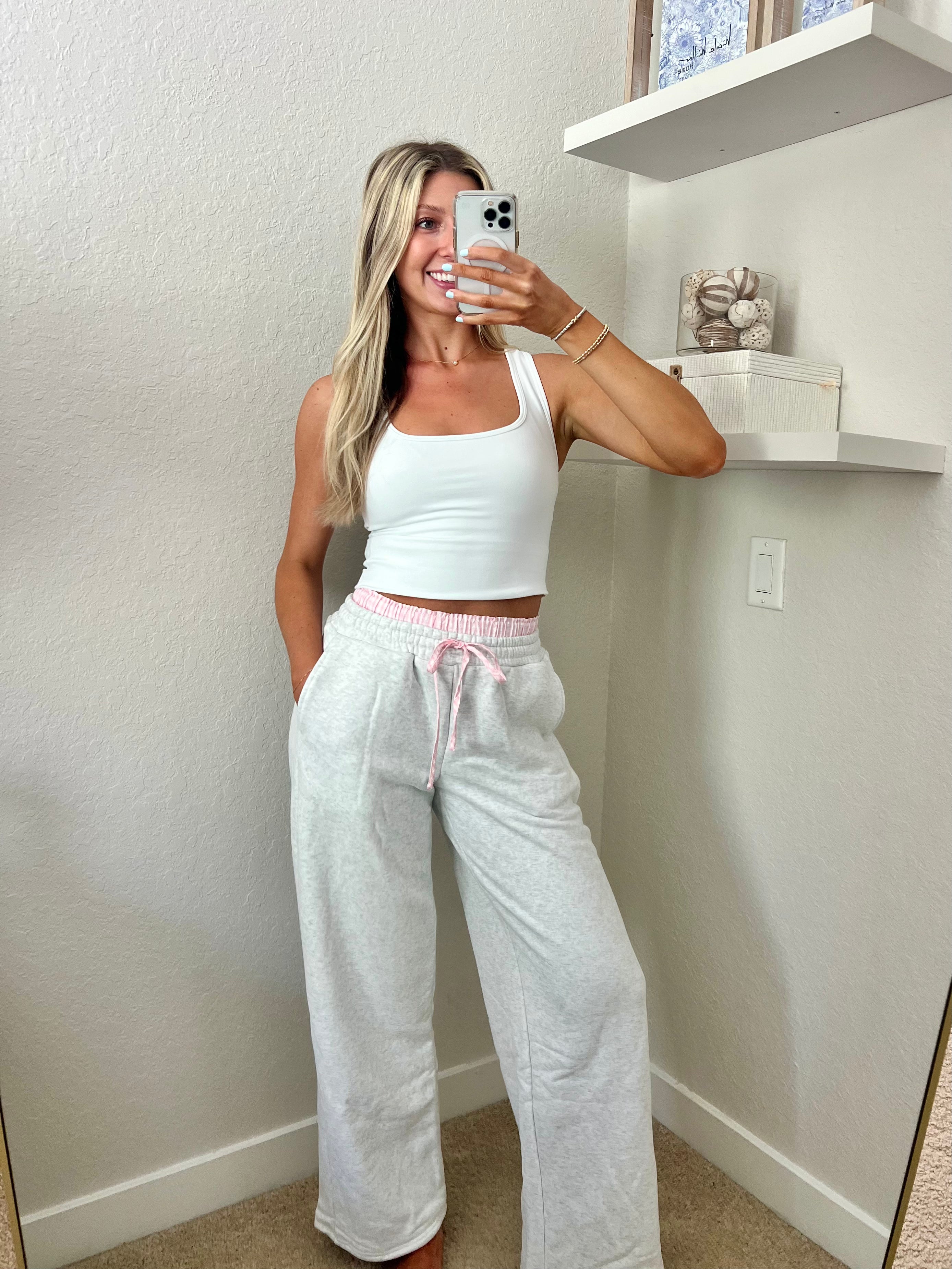 Woman taking a mirror selfie wearing a white tank top and light gray sweatpants with pink and white boxers underneath in a room.