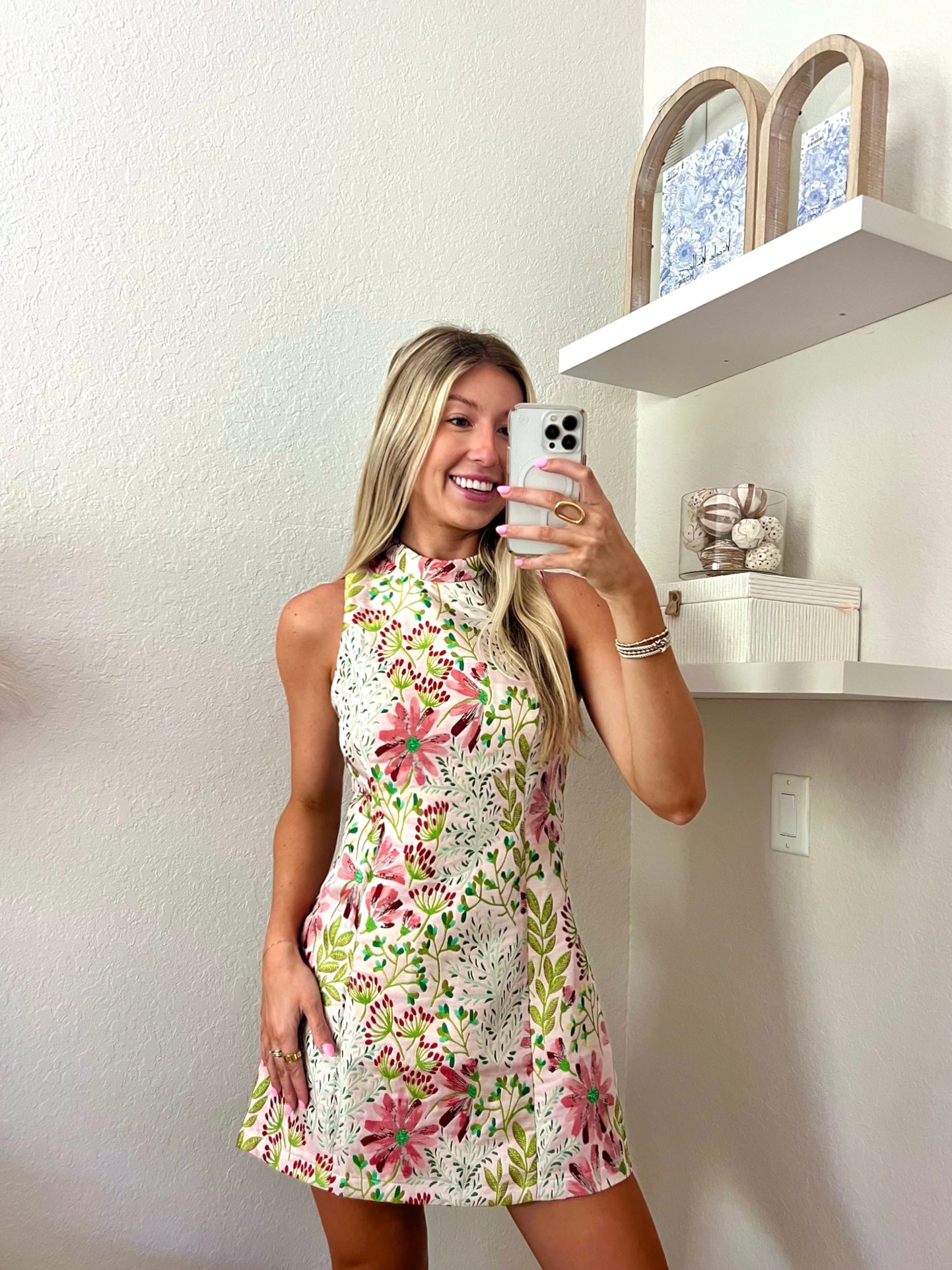 Woman taking a mirror selfie wearing a floral high neck mini dress in a minimalistic room.