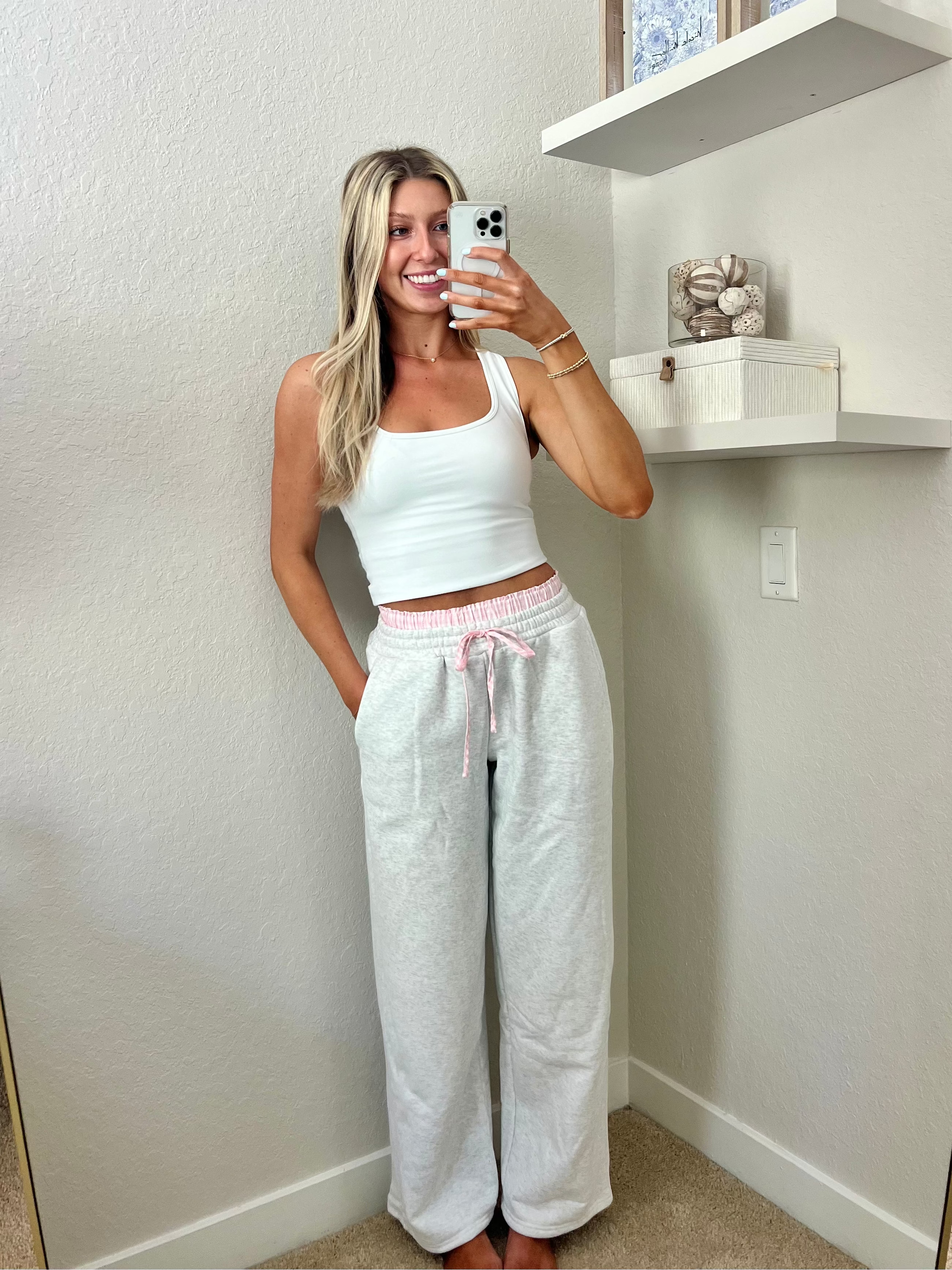 Woman taking a mirror selfie wearing a white tank top and light gray sweatpants with pink and white boxers underneath in a room.