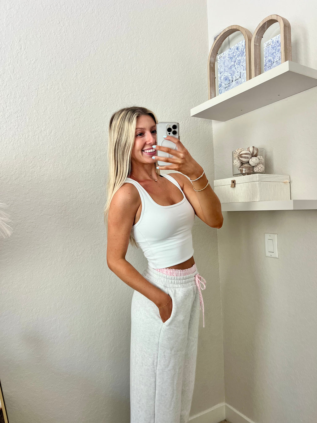 Woman taking a mirror selfie wearing a white tank top and light gray sweatpants with pink and white boxers underneath in a room.