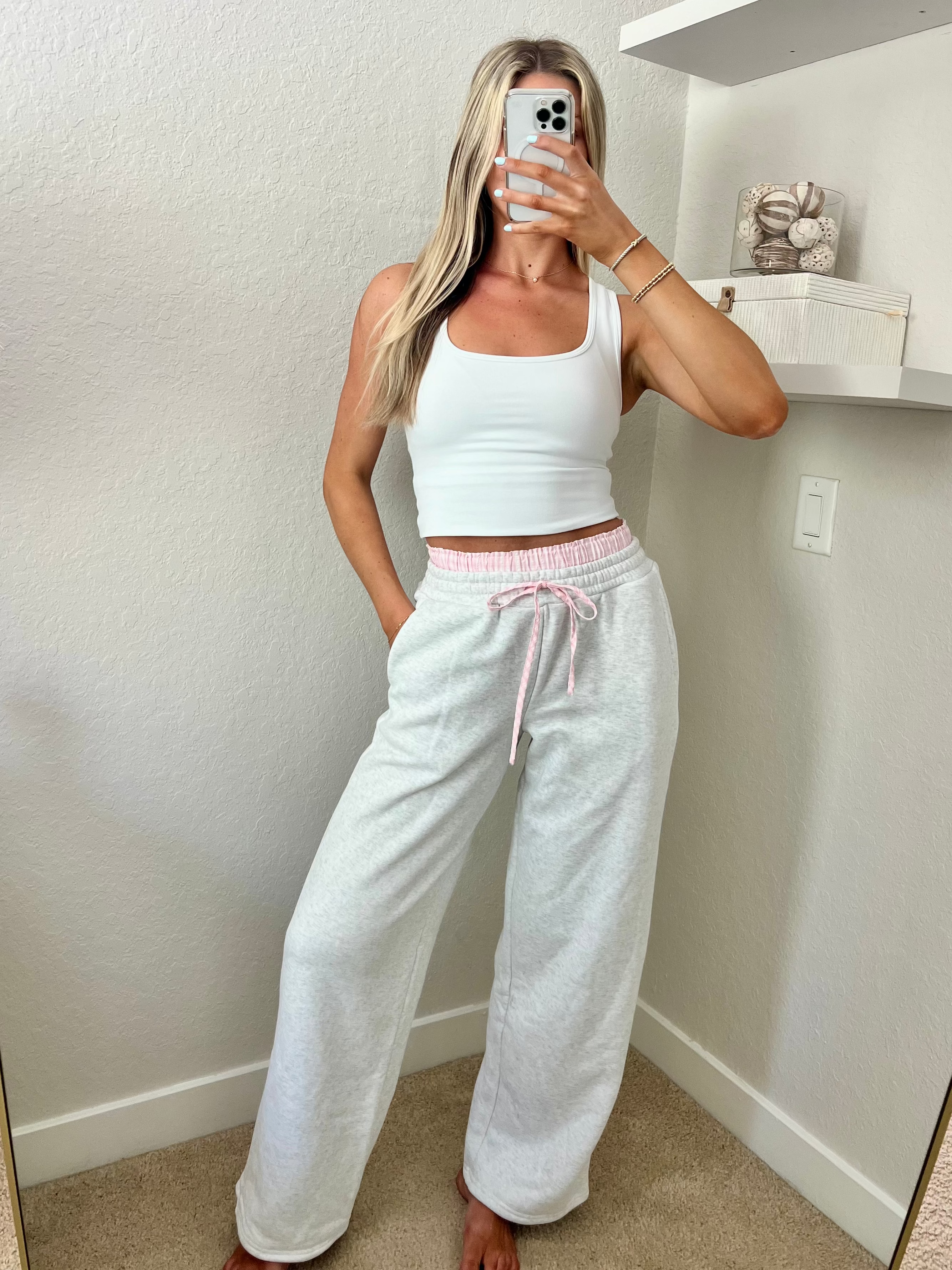 Woman taking a mirror selfie wearing a white tank top and light gray sweatpants with pink and white boxers underneath in a room.