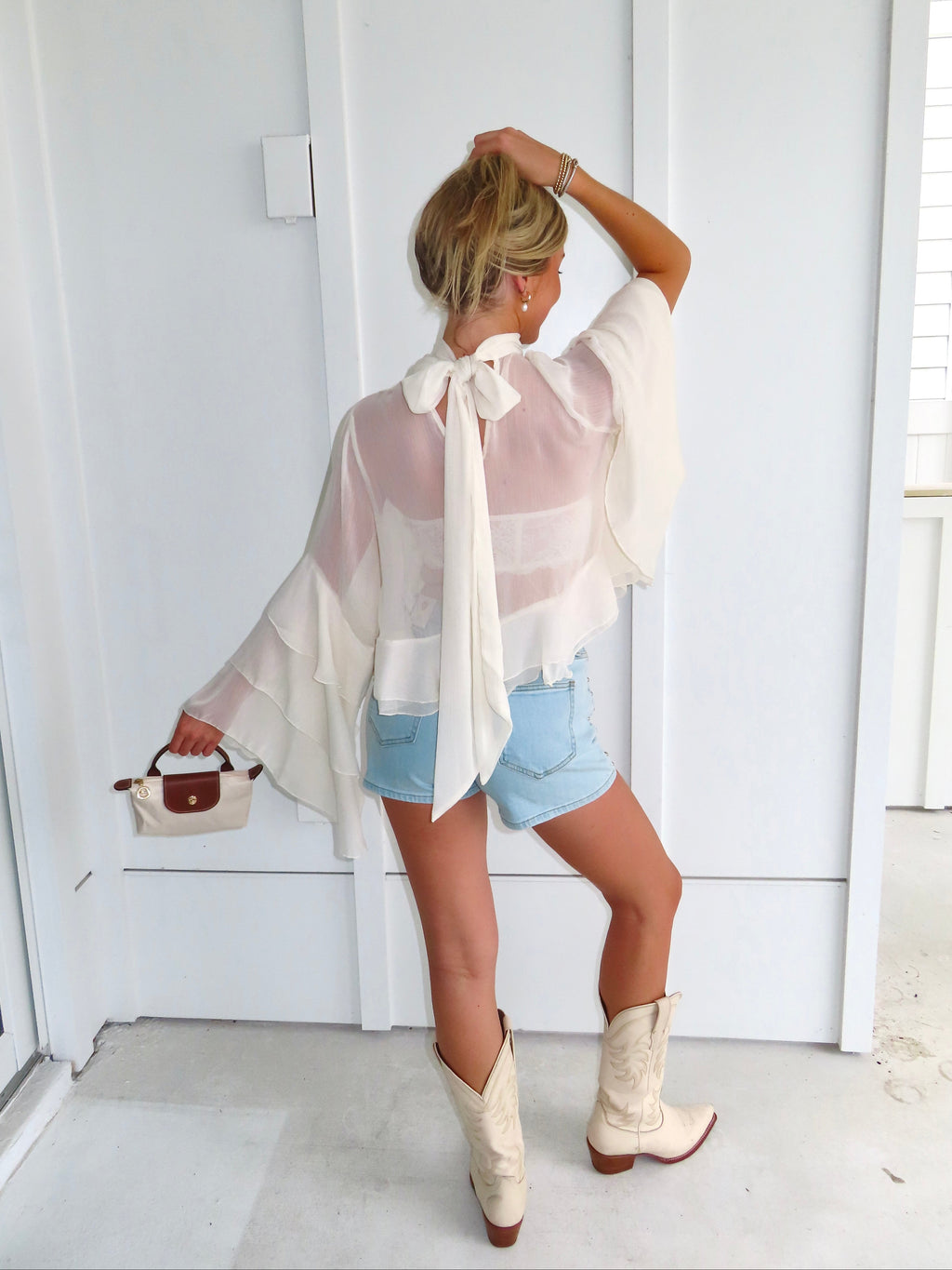 Woman wearing a sheer ivory top, denim shorts, and white cowboy boots in front of a white wall.