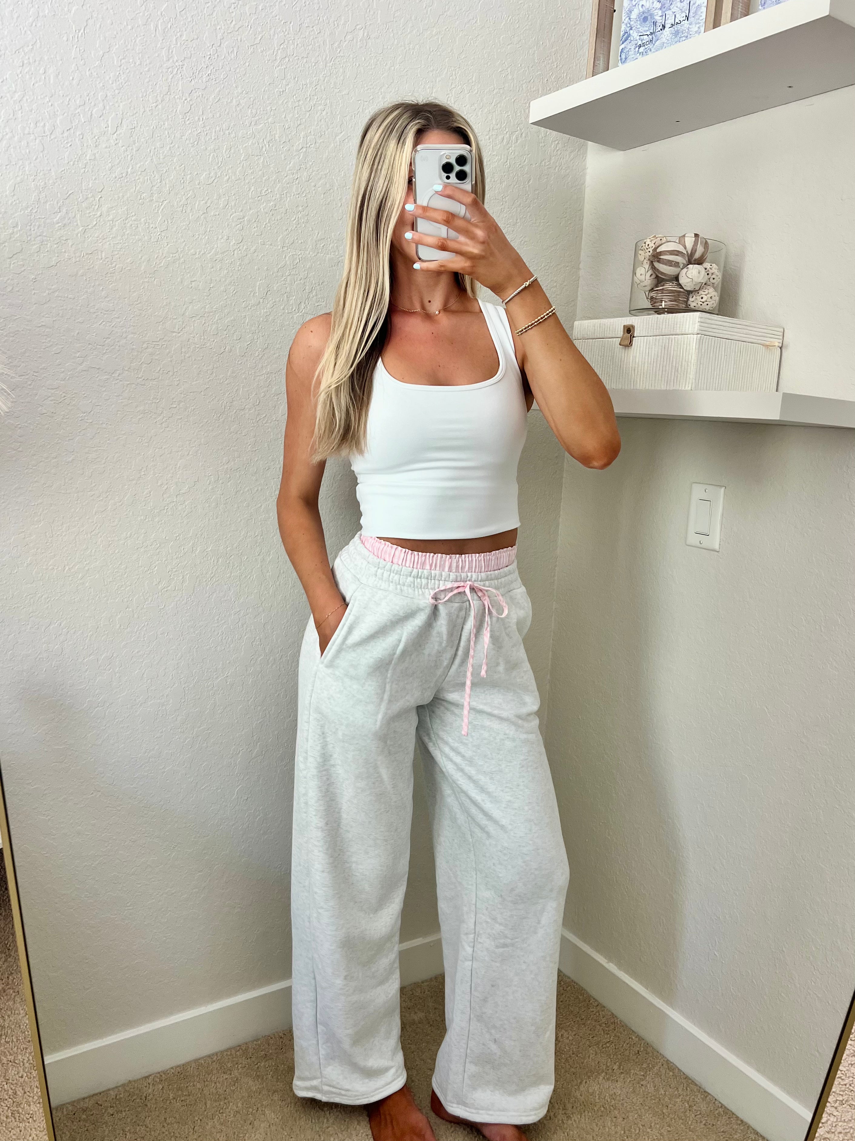 Woman taking a mirror selfie wearing a white tank top and light gray sweatpants with pink and white boxers underneath in a room.