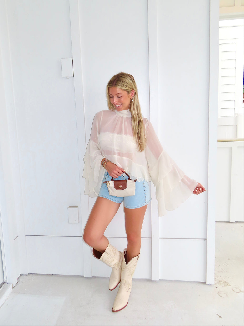 Woman wearing a sheer ivory top, denim shorts, and white cowboy boots, holding a purse in front of a white wall.
