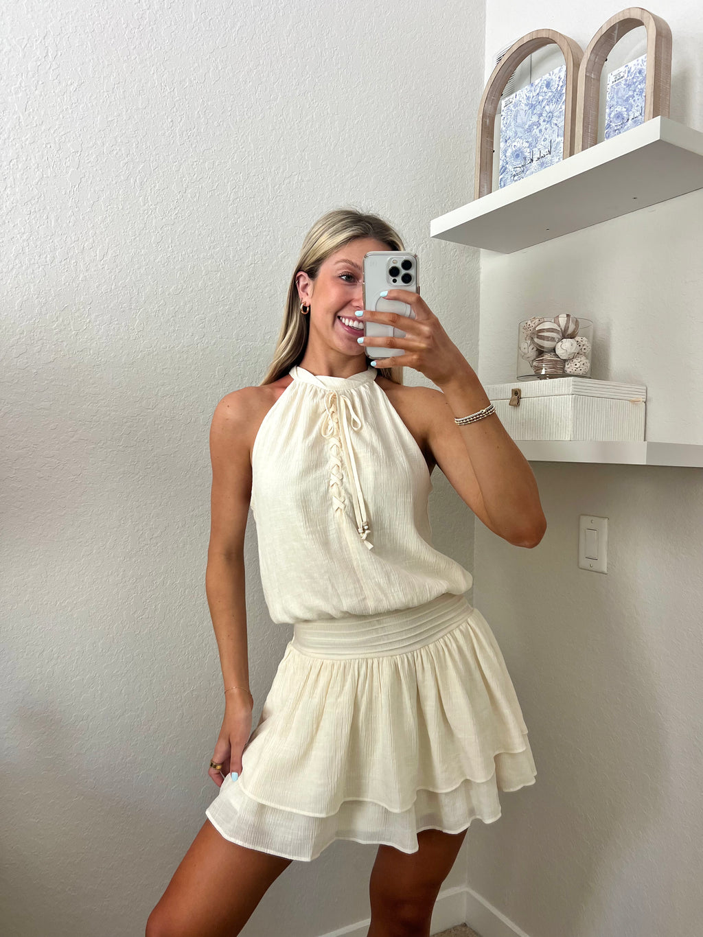 Woman taking a mirror selfie wearing an ivory high neck dress in a room with a beige wall and shelf.