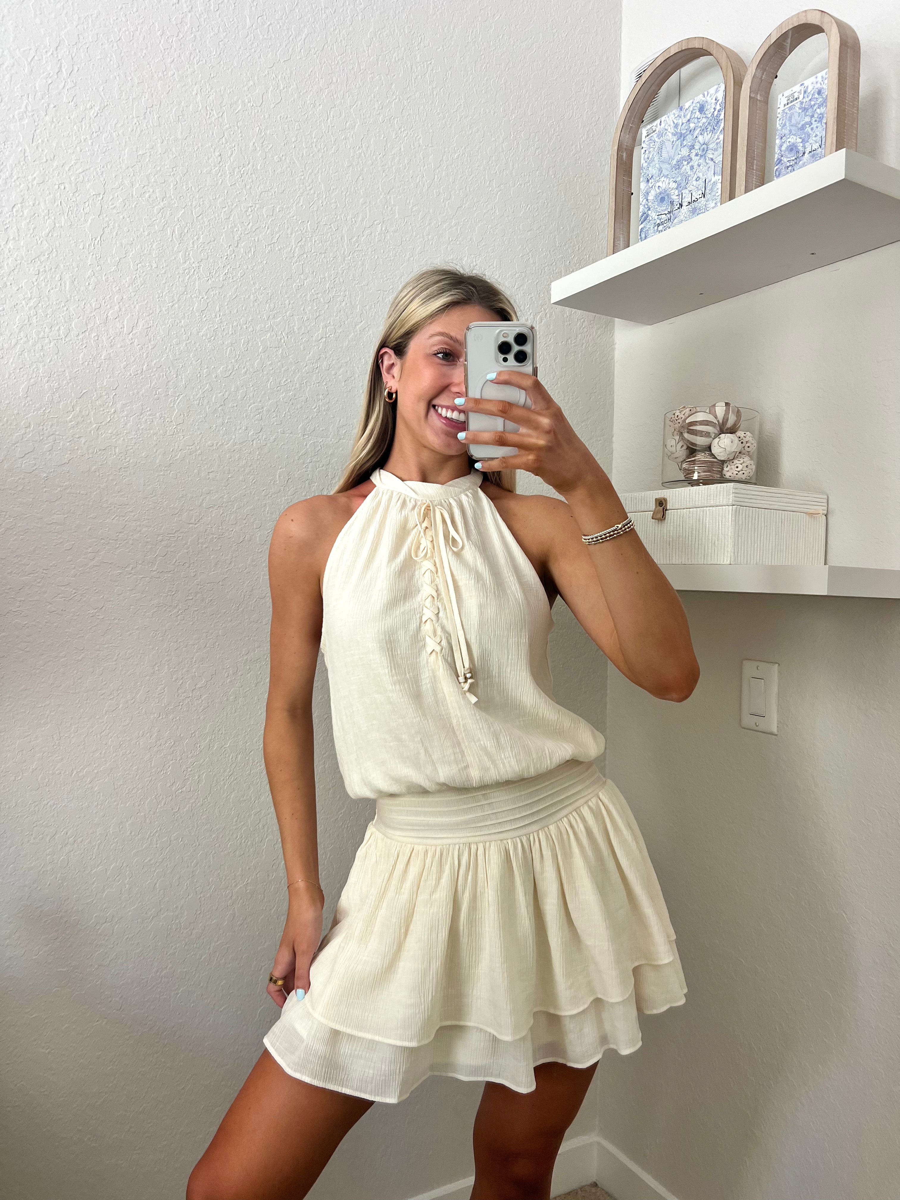 Woman taking a mirror selfie wearing an ivory high neck dress in a room with a beige wall and shelf.