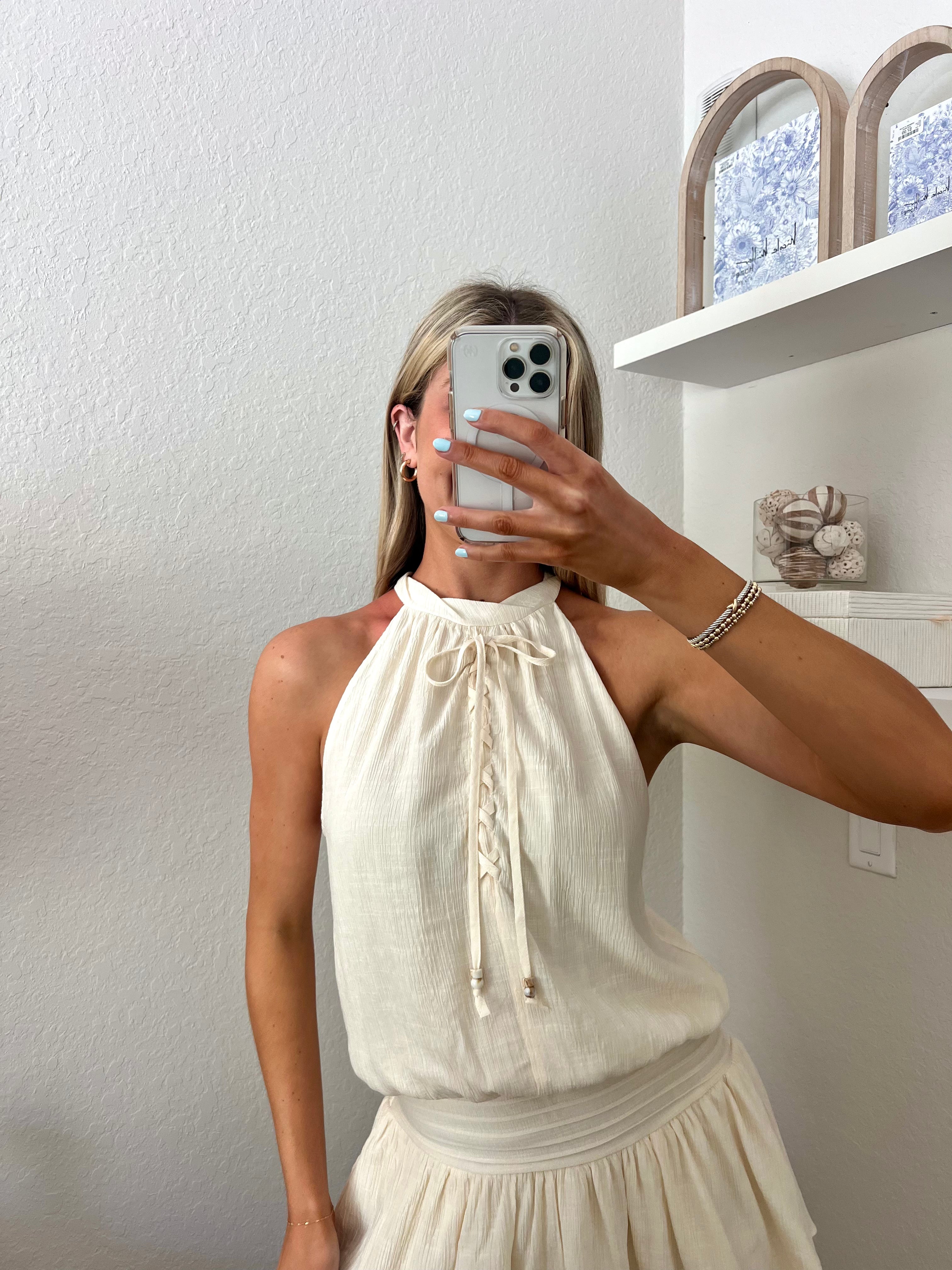 Woman taking a mirror selfie wearing an ivory high neck dress in a room with a beige wall and shelf.