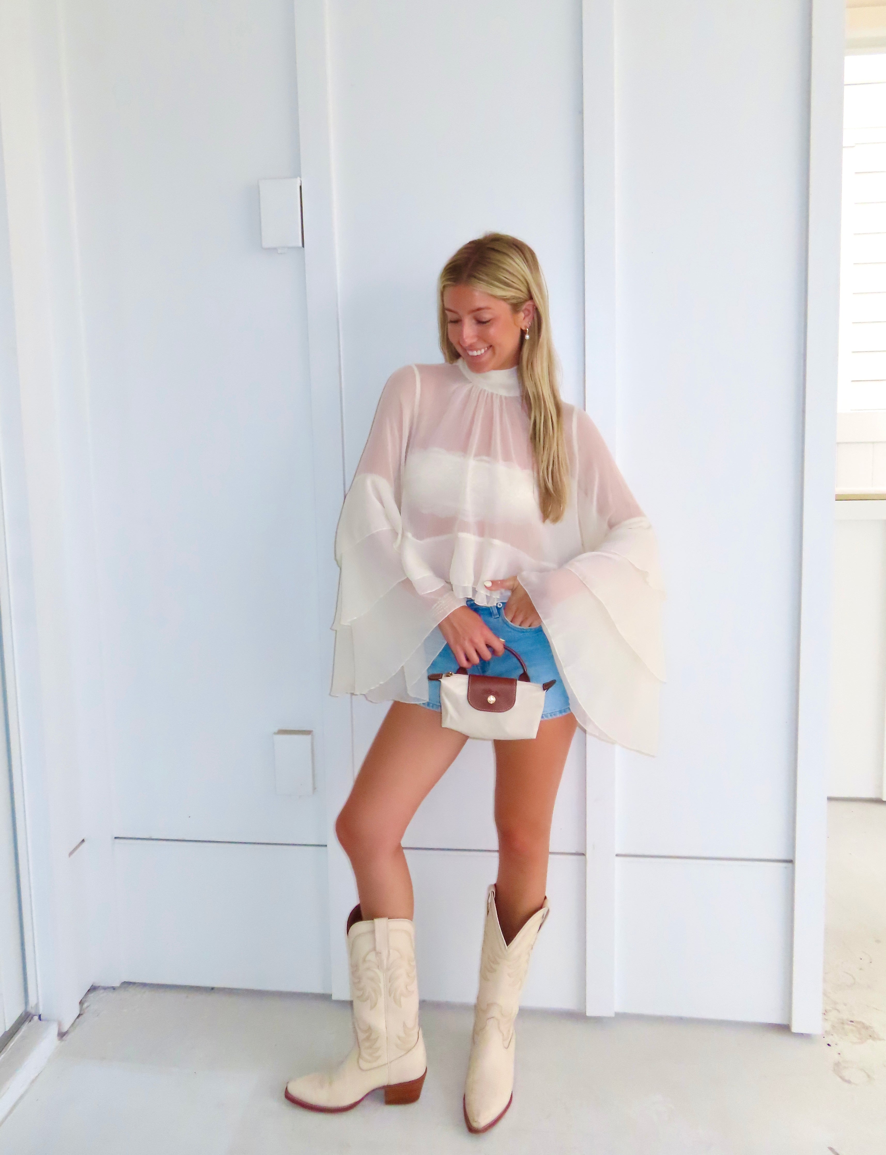 Woman wearing a sheer ivory top, denim shorts, and white cowboy boots, holding a purse in front of a white wall.