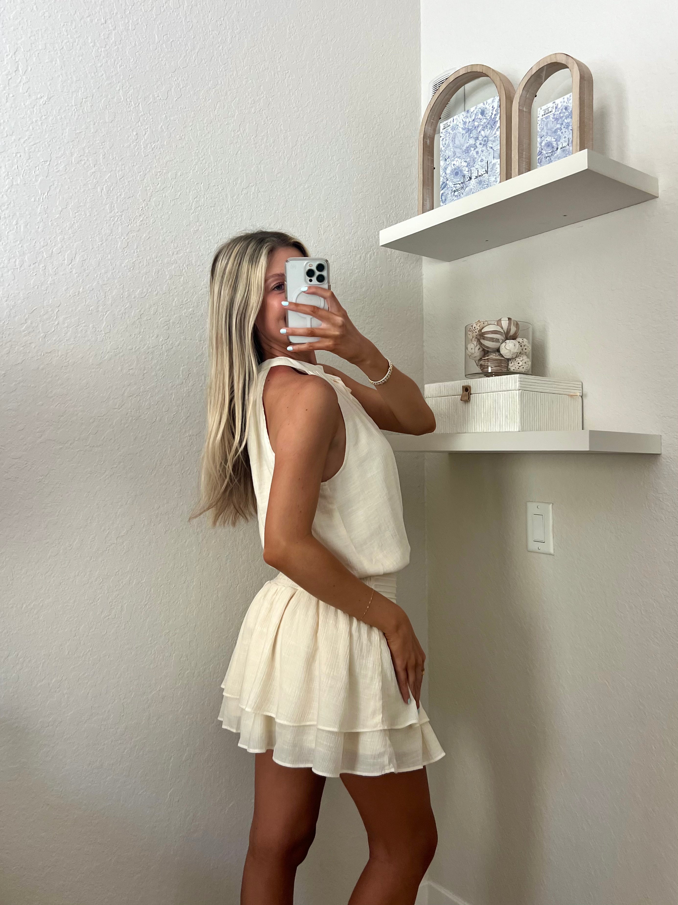 Woman taking a mirror selfie wearing an ivory high neck dress in a room with a beige wall and shelf.