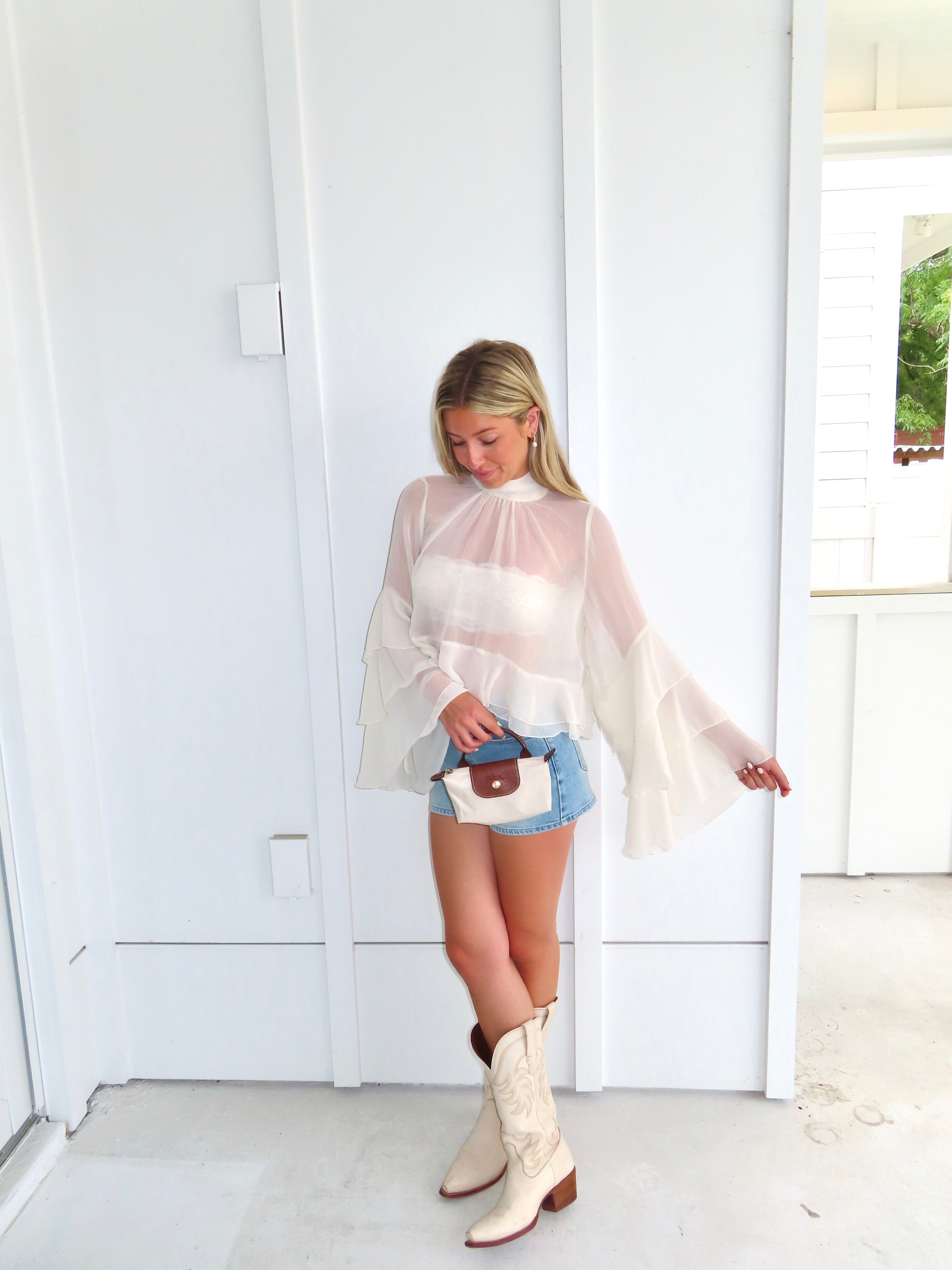 Woman wearing a sheer ivory top, denim shorts, and white cowboy boots, holding a purse in front of a white wall.