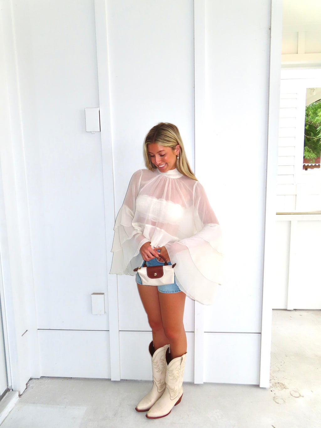 Woman wearing a sheer ivory top, denim shorts, and white cowboy boots, holding a purse in front of a white wall.