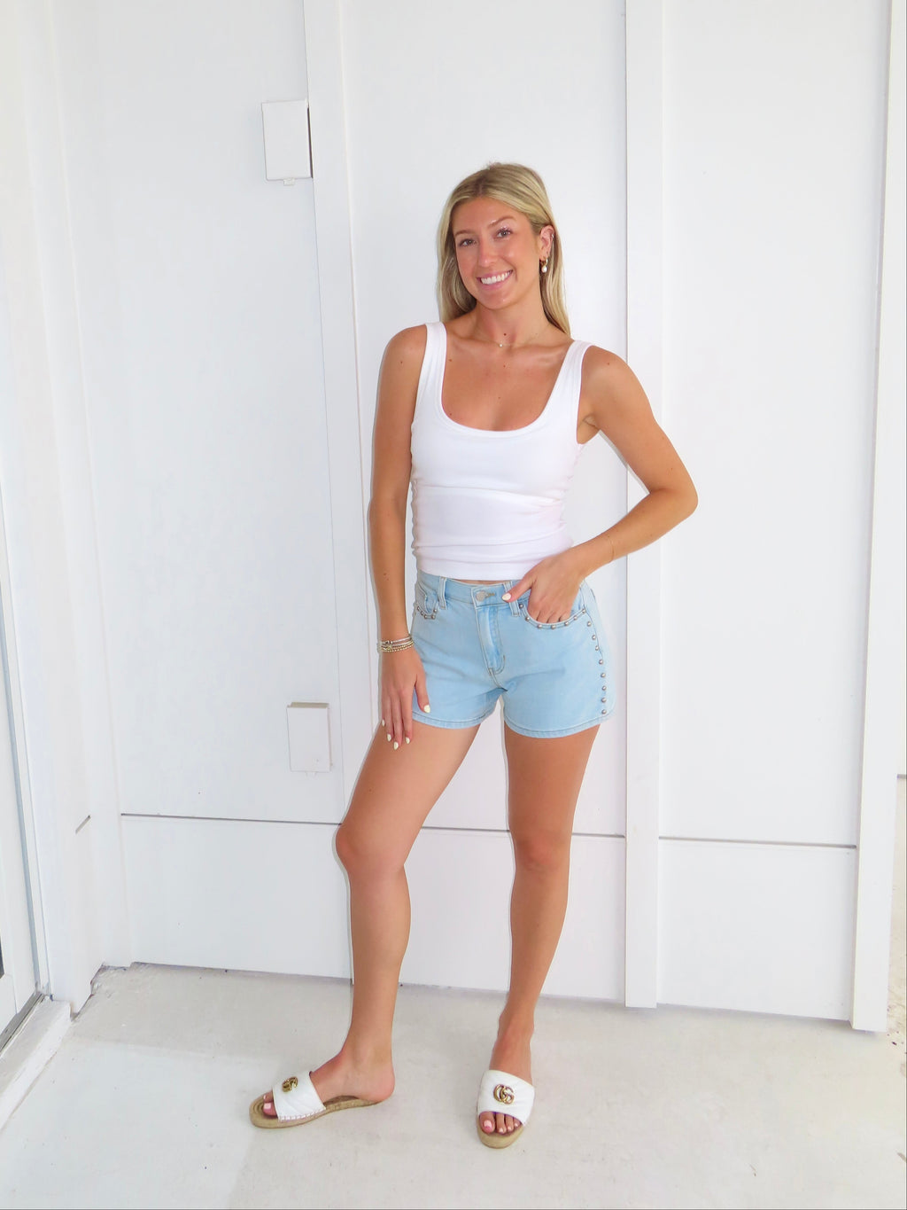 Woman wearing a white tank top and light blue denim shorts standing against a white wall.