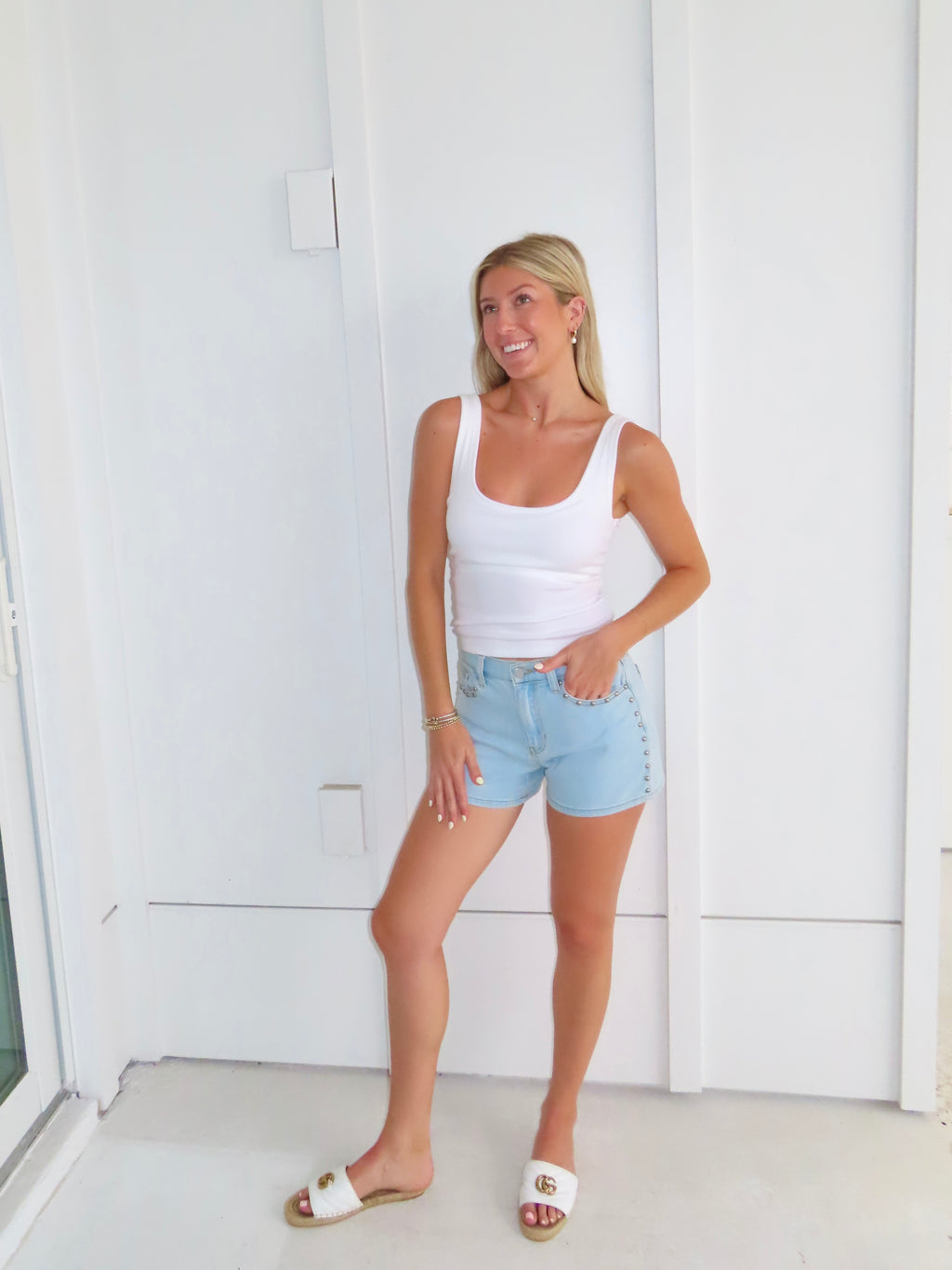 Woman wearing a white tank top and light blue denim shorts standing against a white wall.
