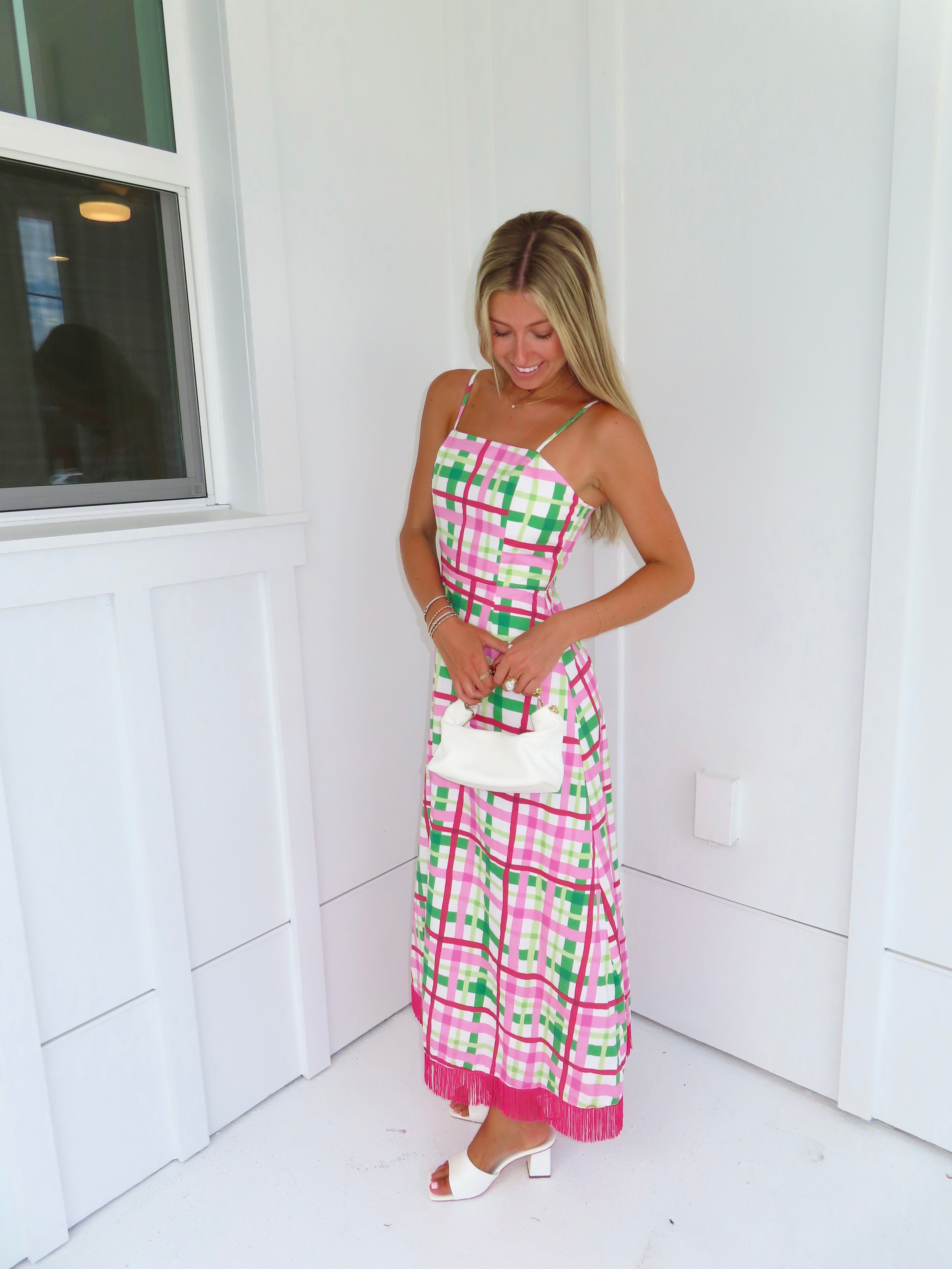 Woman wearing a pink and green plaid maxi dress holding a white purse against a minimal white wall.