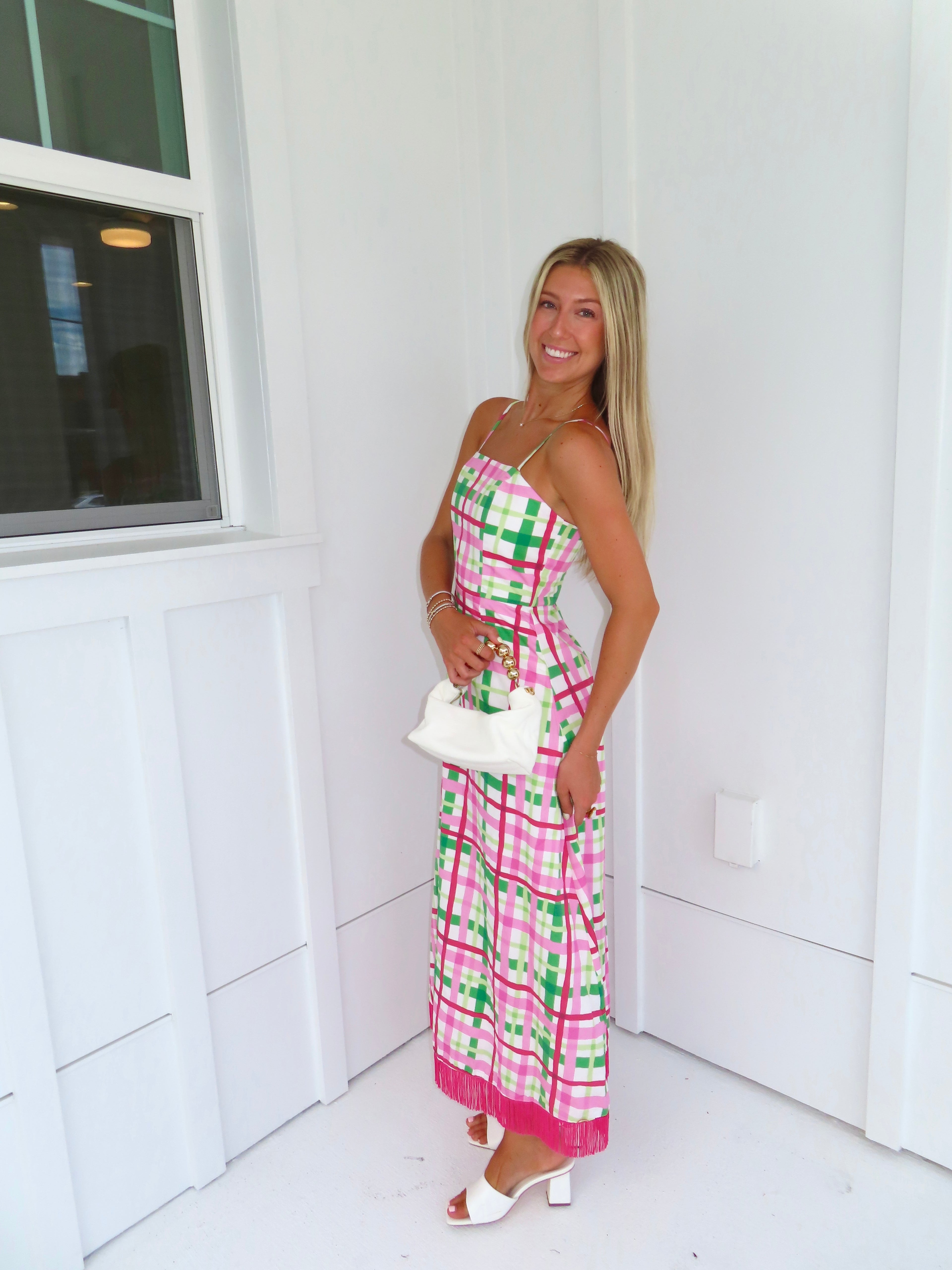 Woman wearing a pink and green plaid maxi dress holding a white purse against a minimal white wall.