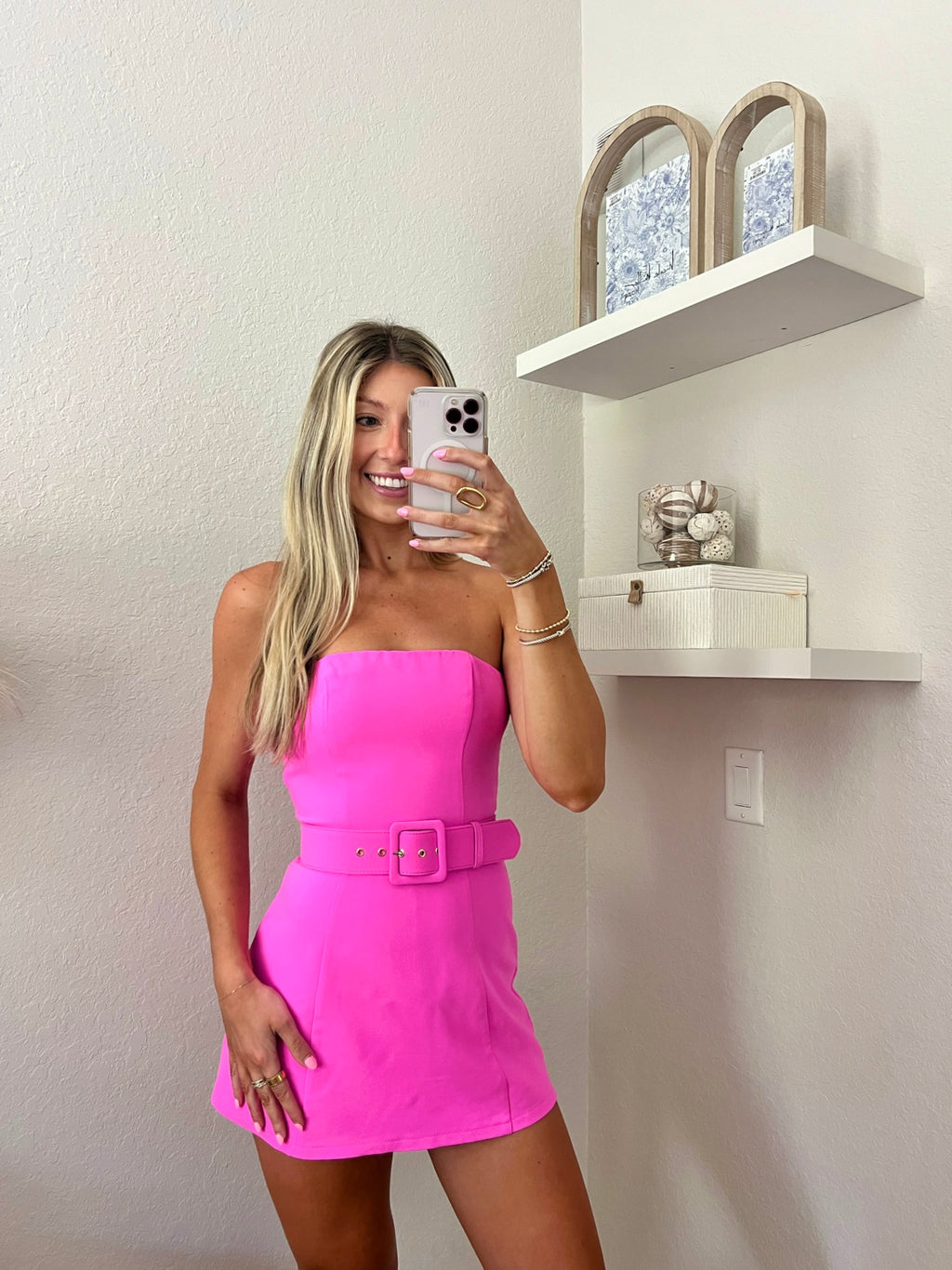 Woman taking a mirror selfie wearing a bright pink strapless dress with a belt.