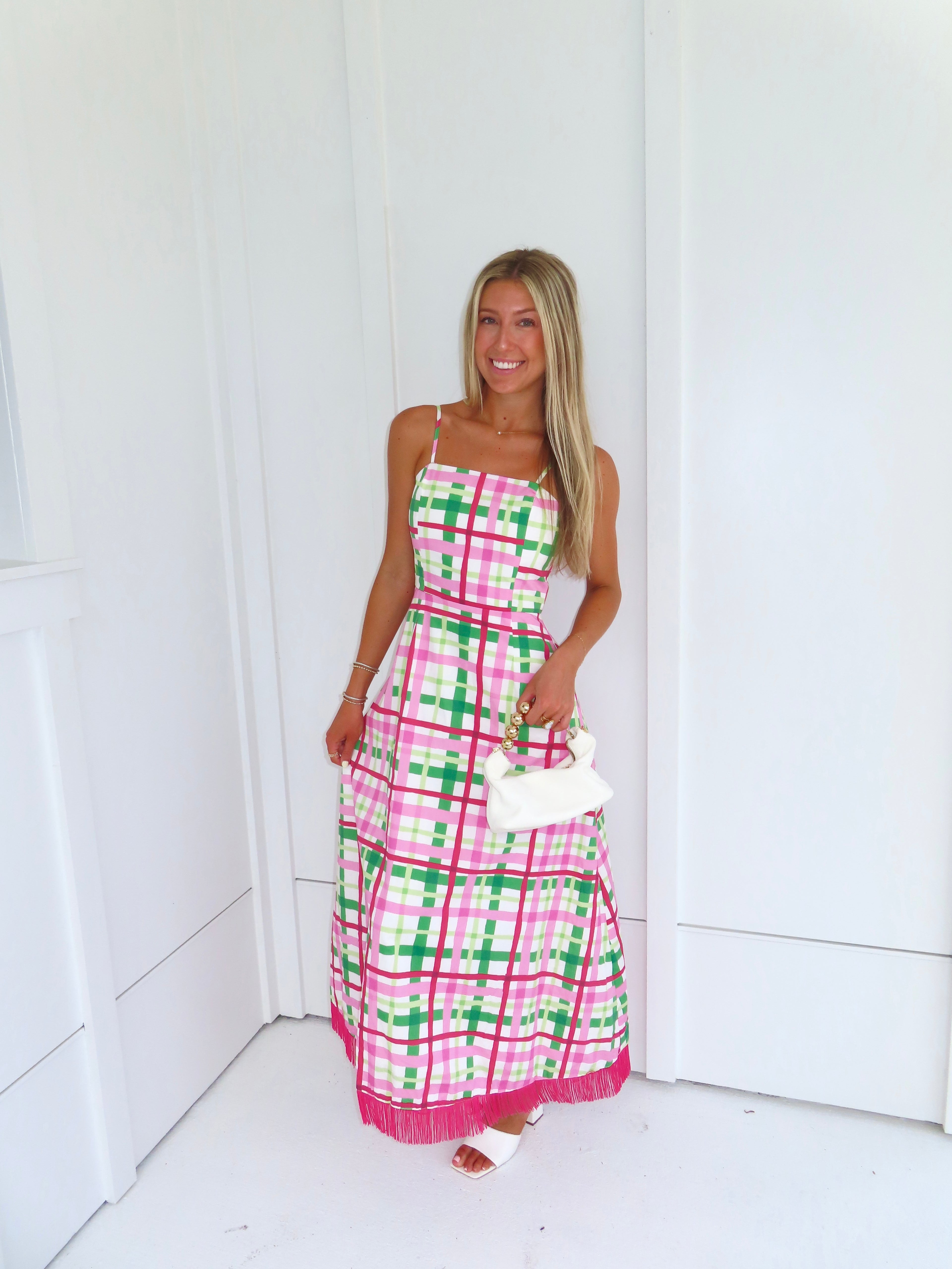 Woman wearing a pink and green plaid maxi dress holding a white purse against a minimal white wall.