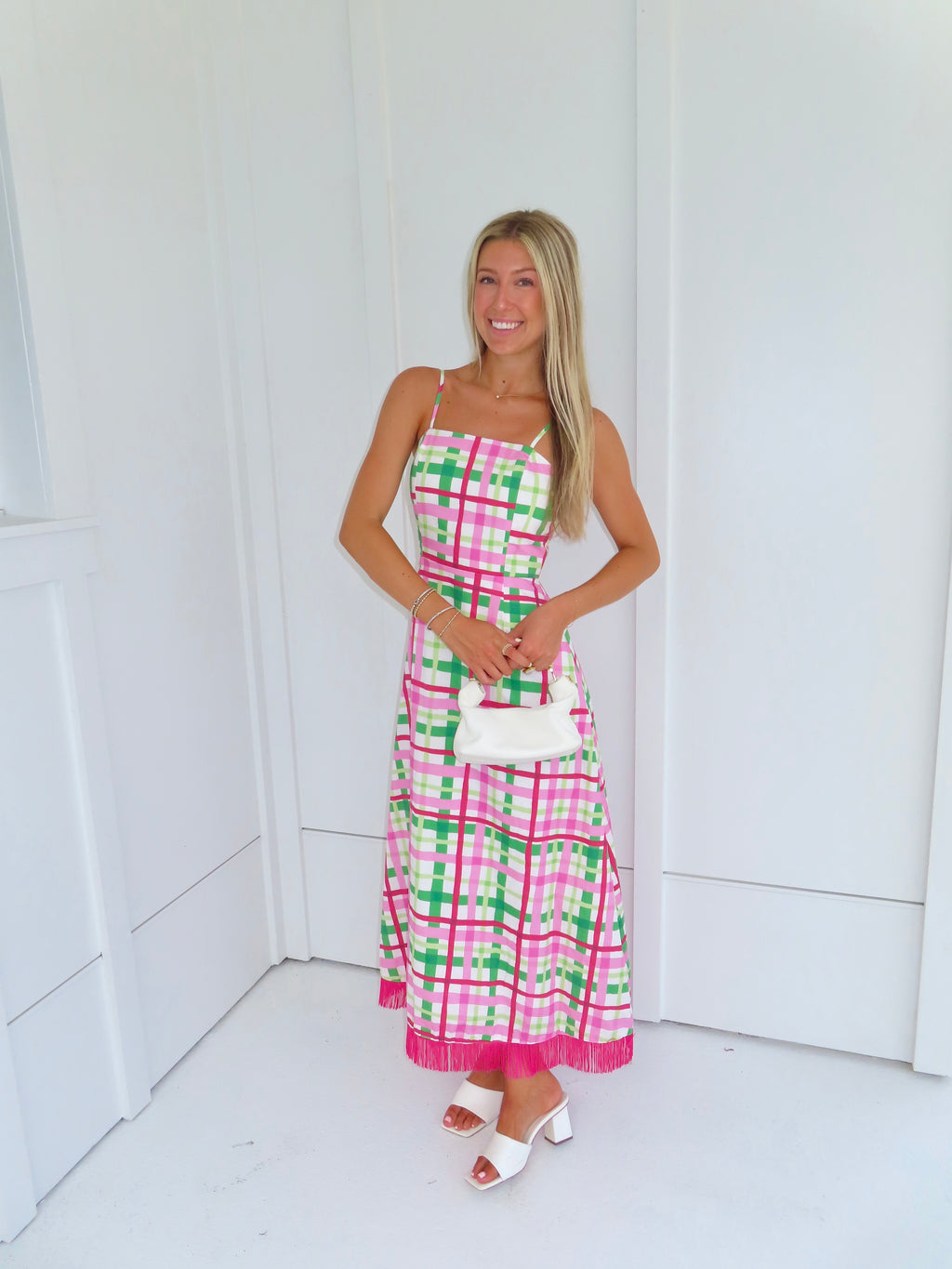 Woman wearing a pink and green plaid maxi dress holding a white purse against a minimal white wall.