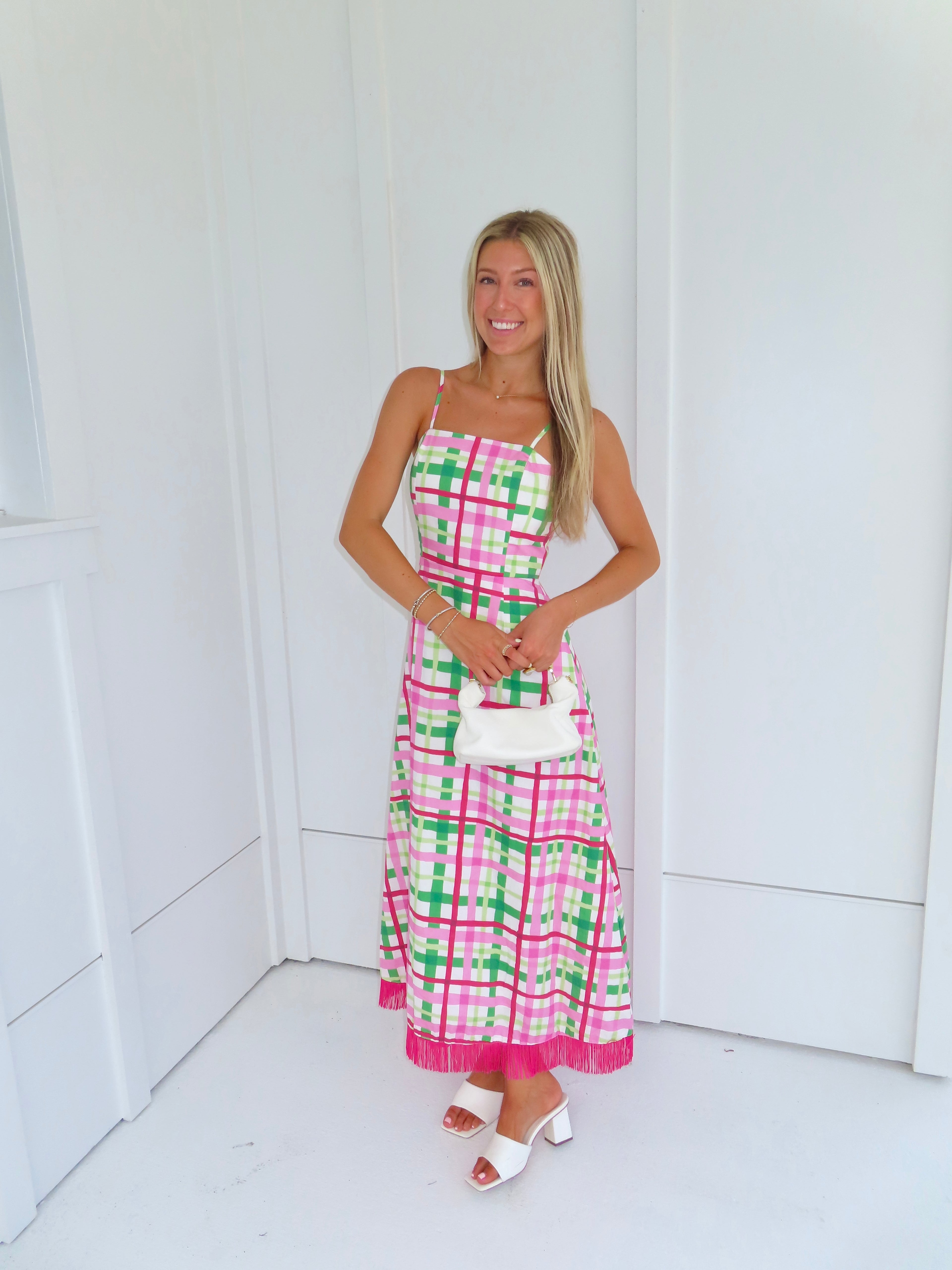 Woman wearing a pink and green plaid maxi dress holding a white purse against a minimal white wall.