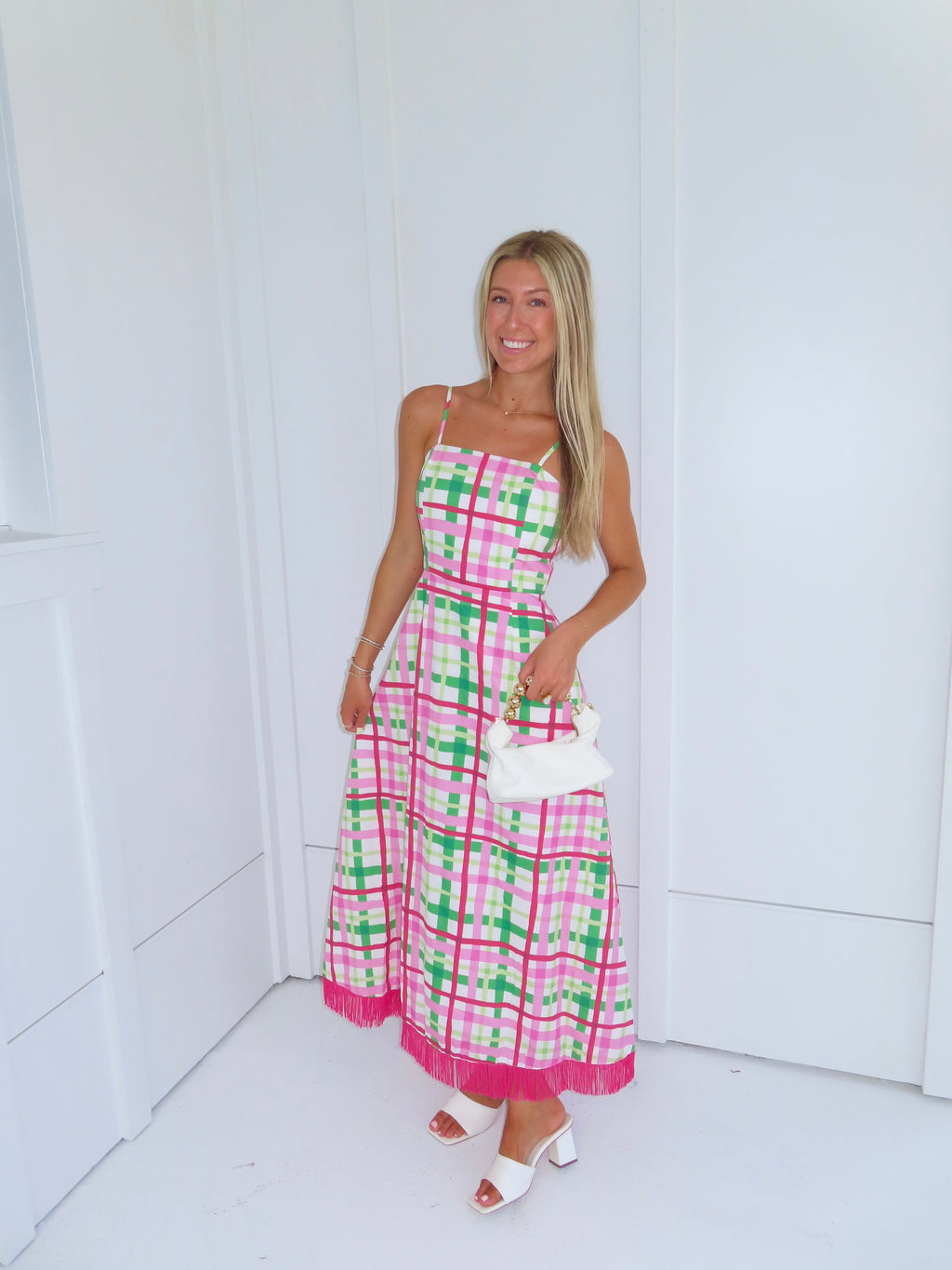 Woman wearing a pink and green plaid maxi dress holding a white purse against a minimal white wall.