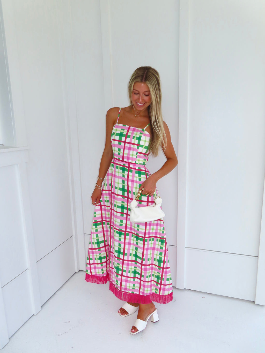 Woman wearing a pink and green plaid maxi dress holding a white purse against a minimal white wall.