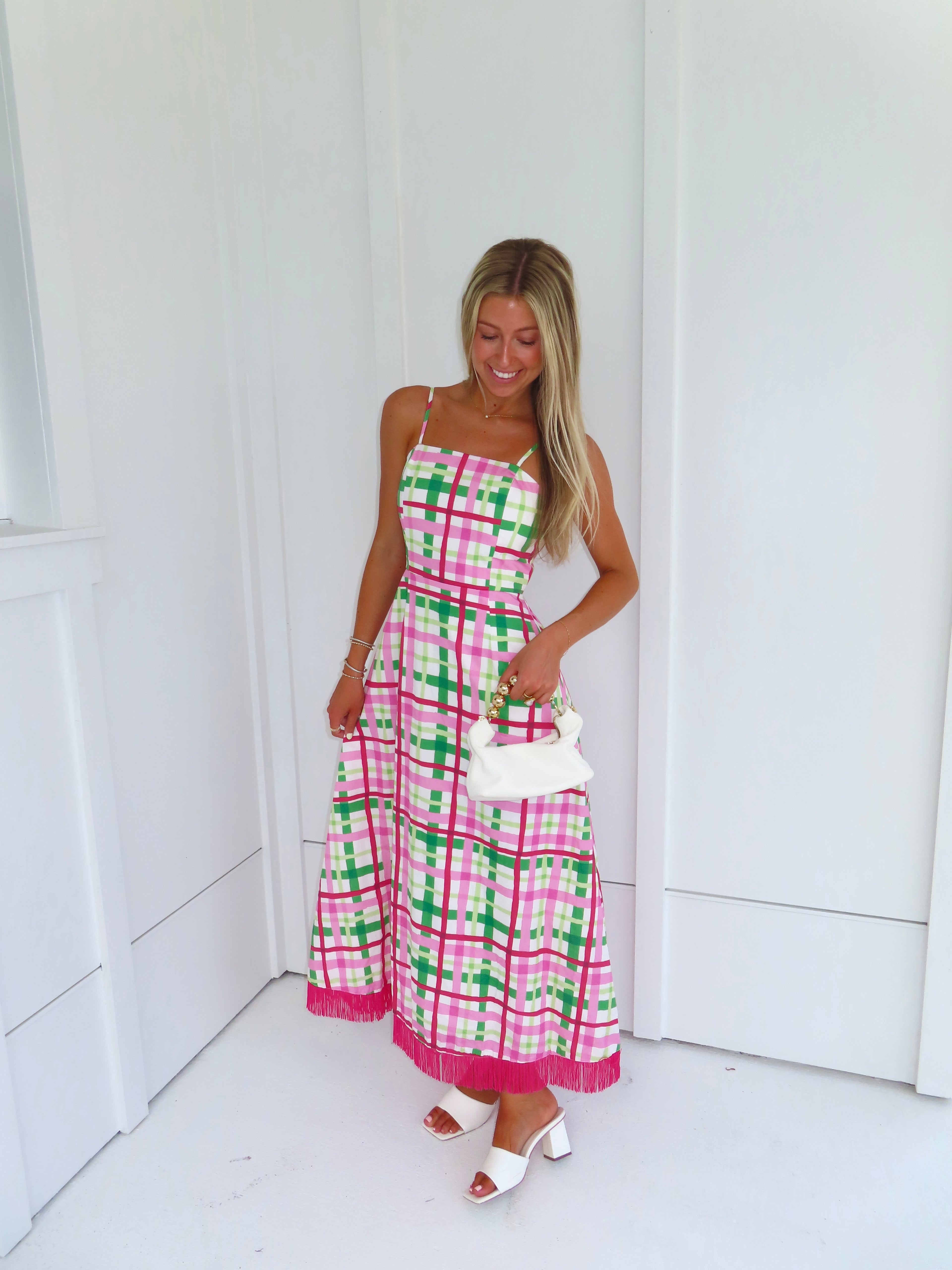 Woman wearing a pink and green plaid maxi dress holding a white purse against a minimal white wall.