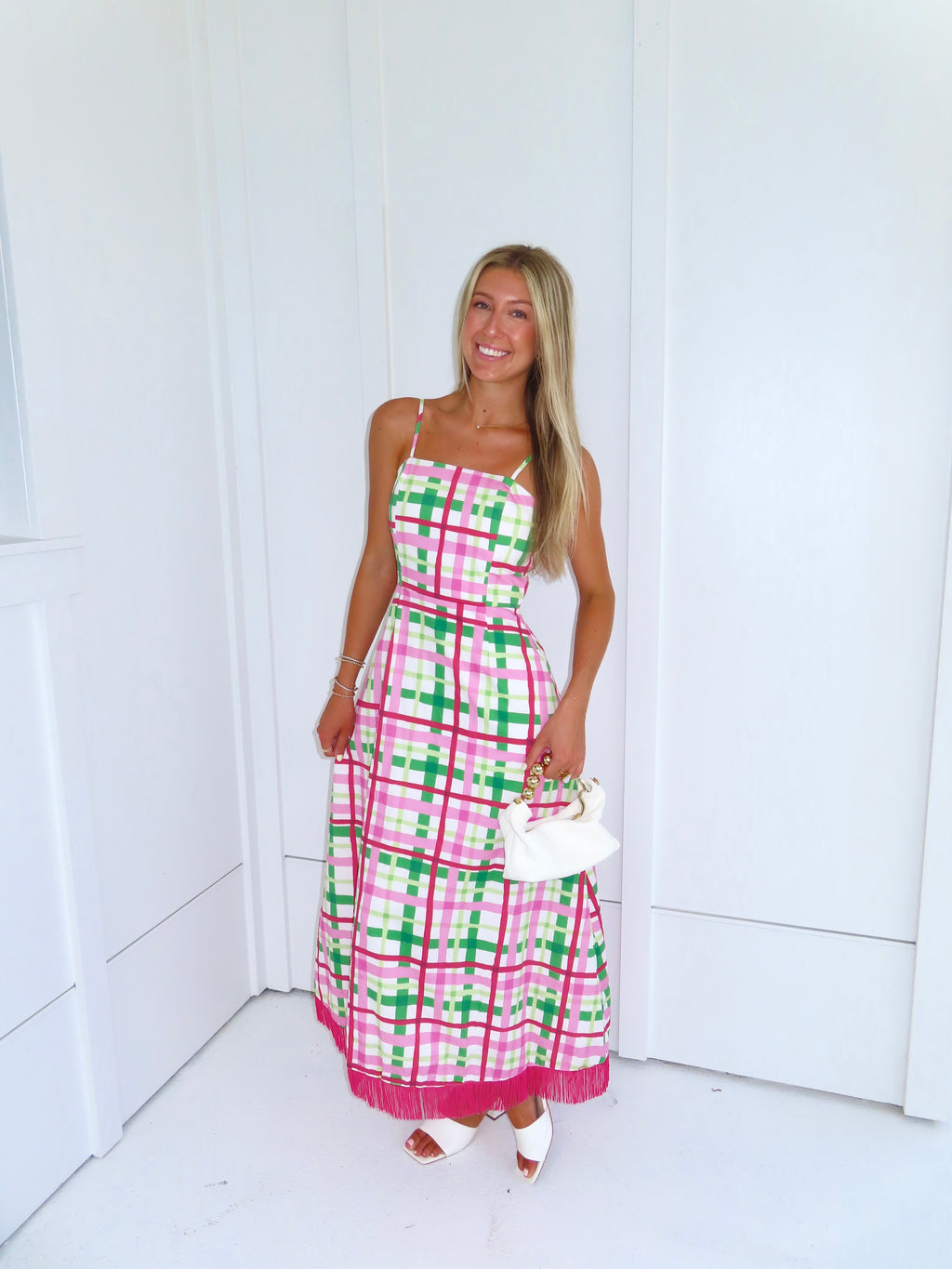 Woman wearing a pink and green plaid maxi dress holding a white purse against a minimal white wall.