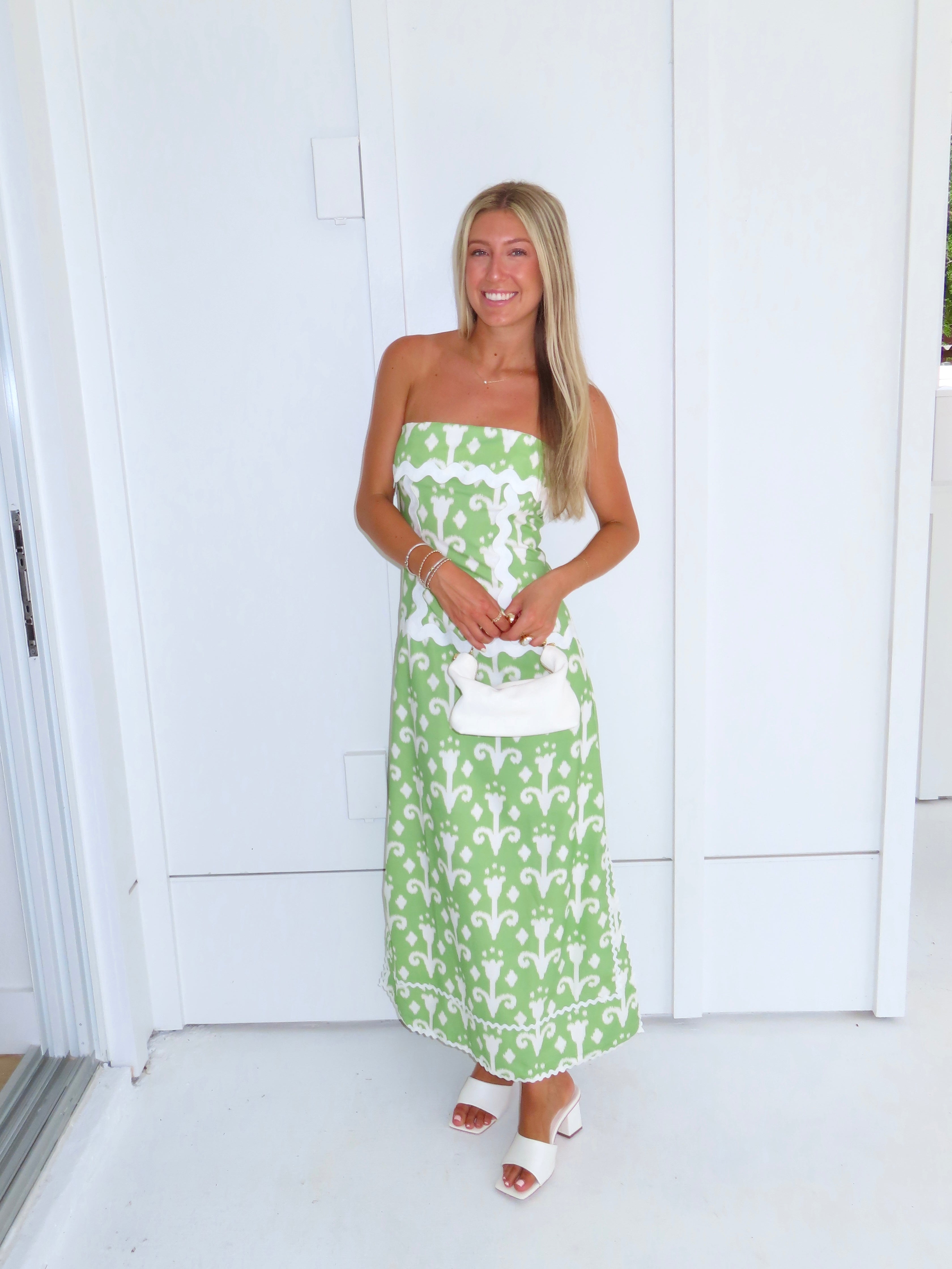 Woman wearing a strapless green printed maxi dress holding a white purse against a white wall.