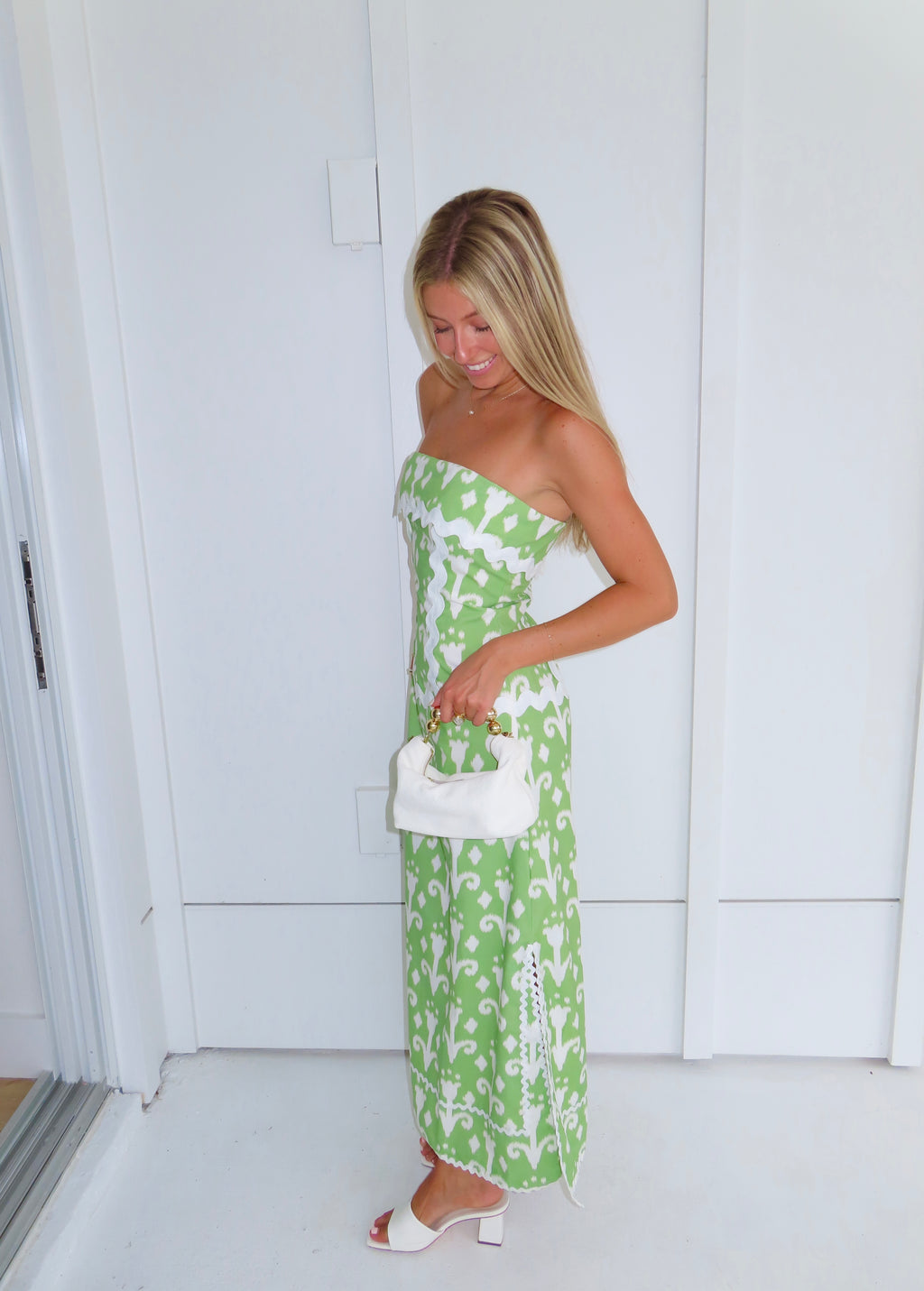 Woman wearing a strapless green printed maxi dress holding a white purse against a white wall.