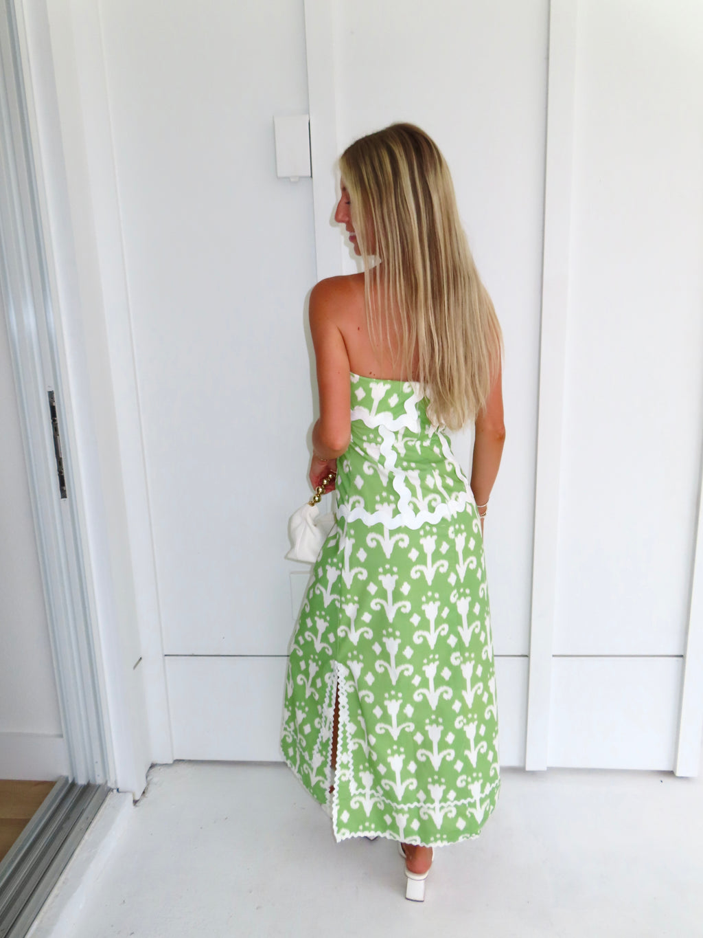 Woman wearing a strapless green printed maxi dress holding a white purse against a white wall.