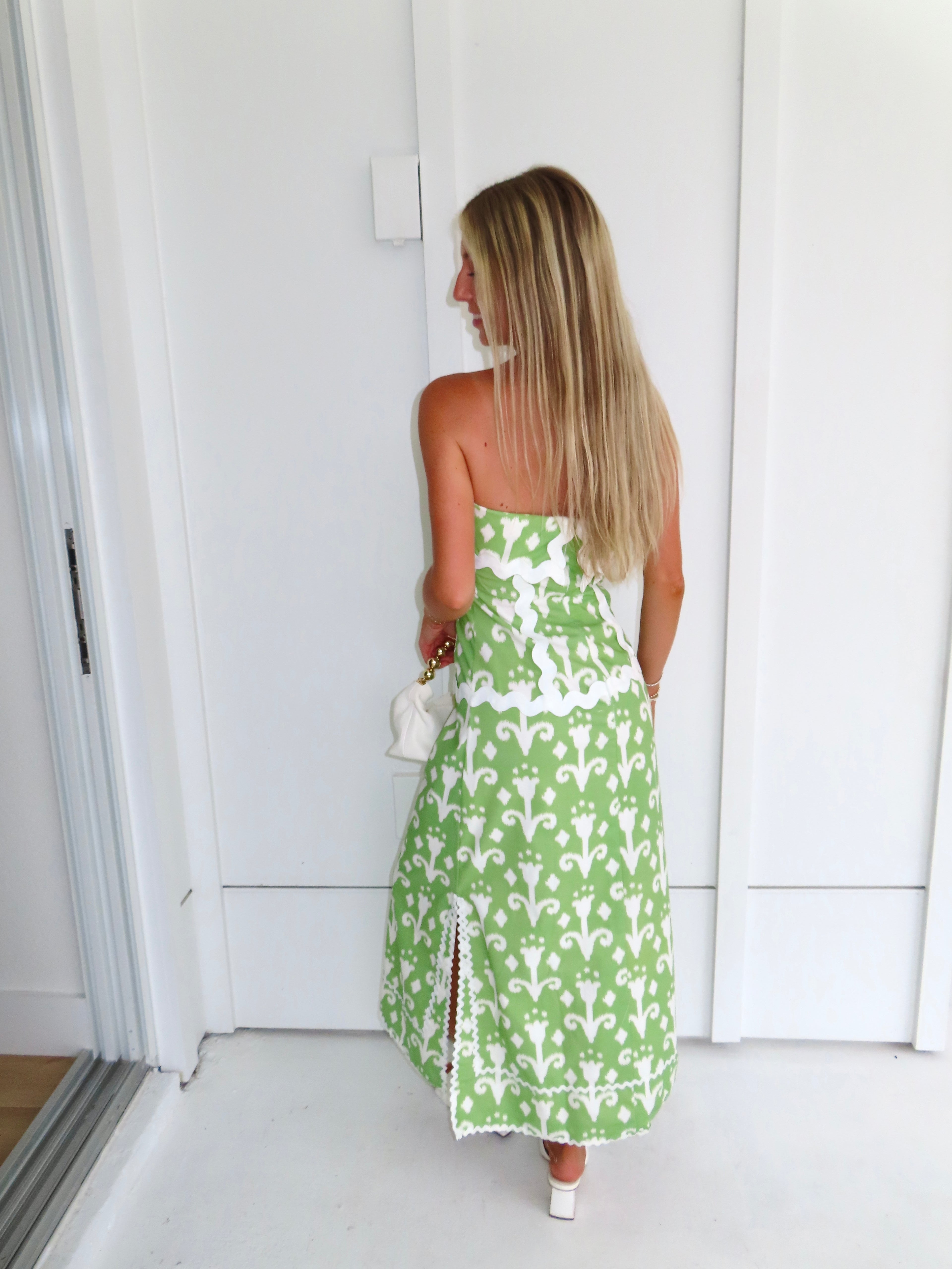 Woman wearing a strapless green printed maxi dress holding a white purse against a white wall.
