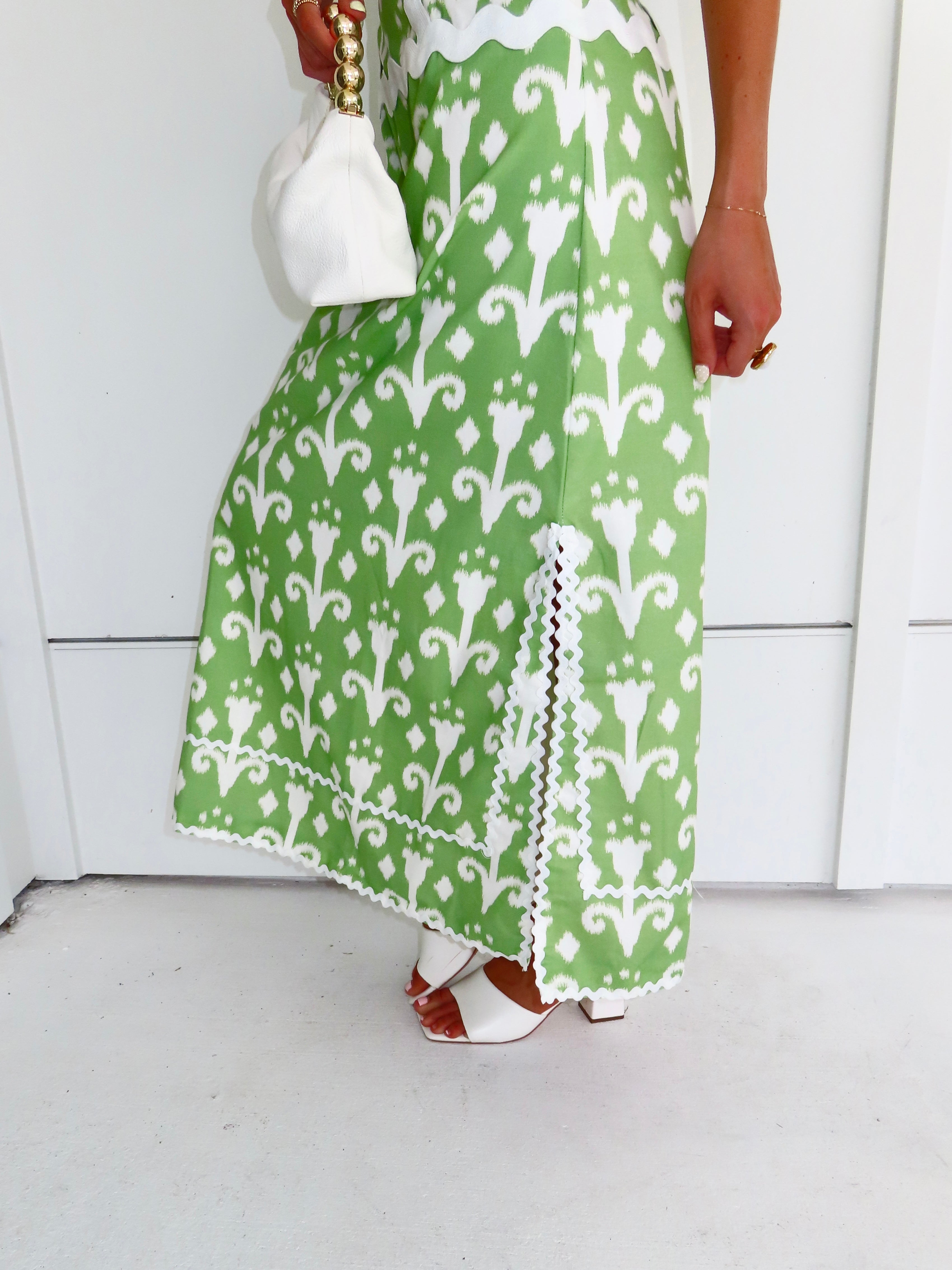Woman wearing a strapless green printed maxi dress holding a white purse against a white wall.