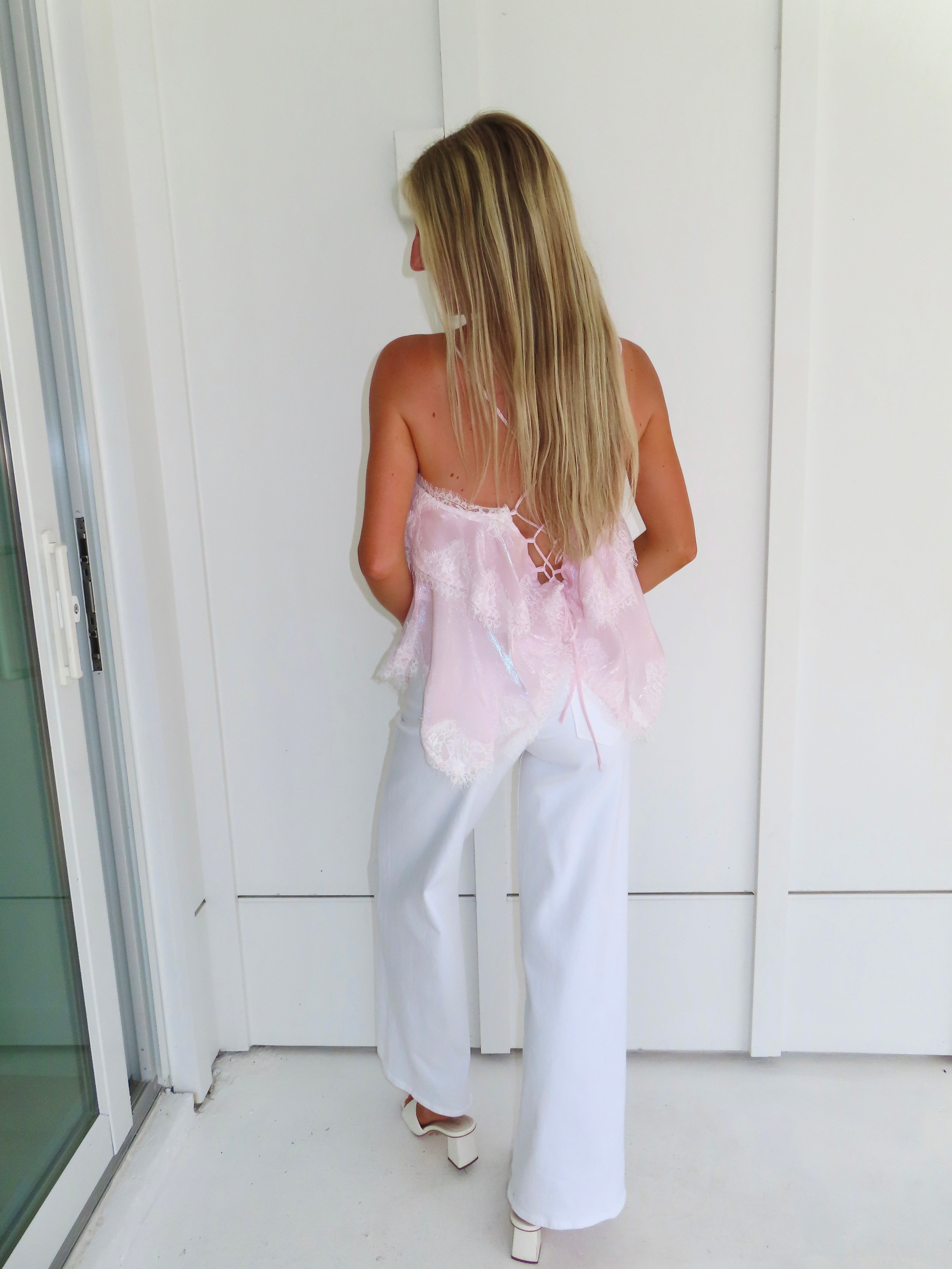 Woman wearing a light pink ruffled tank top and white pants, standing against a white wall.