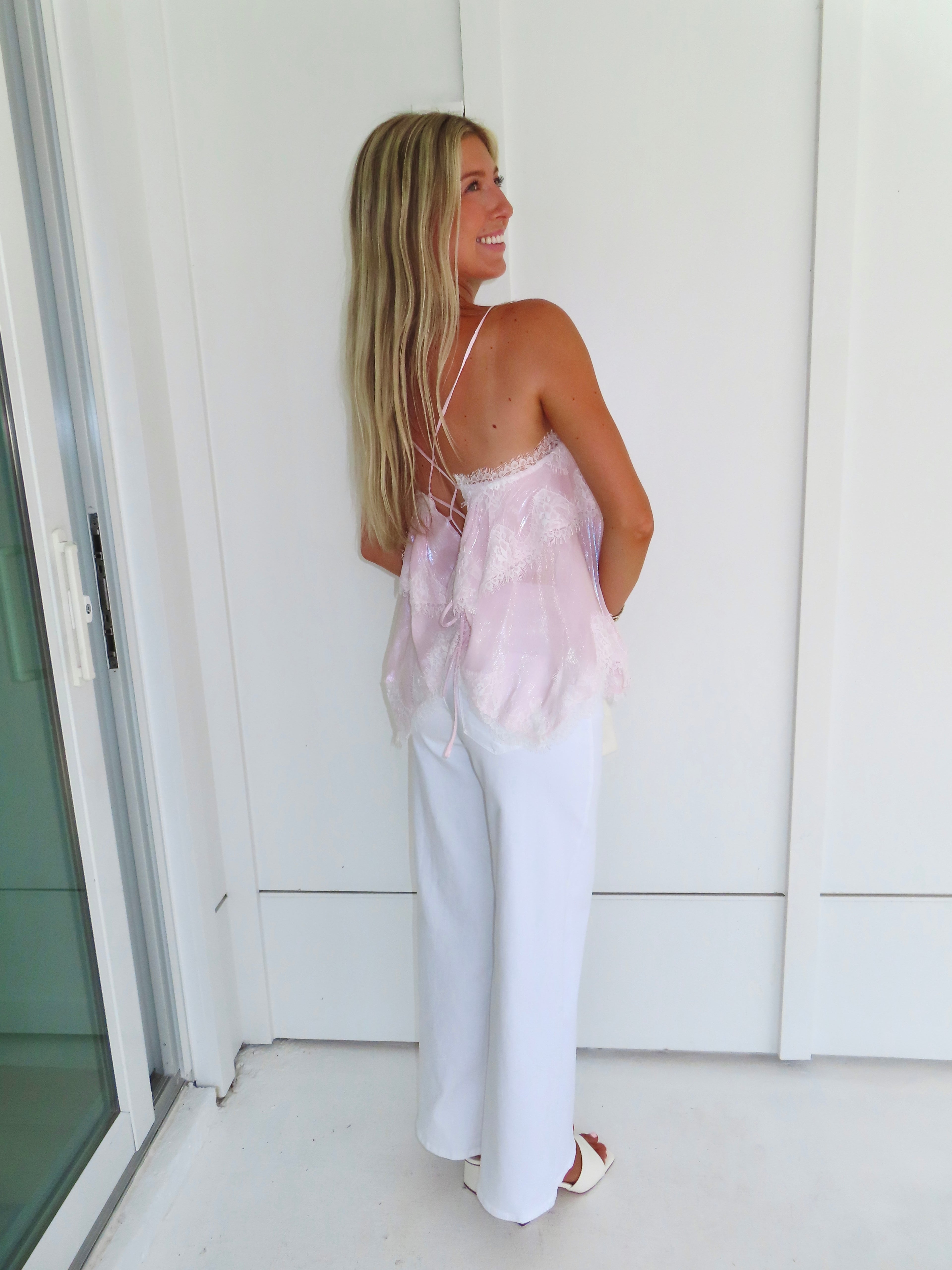 Woman wearing a light pink ruffled tank top and white pants, standing against a white wall.