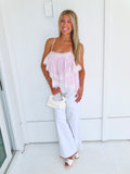 Woman wearing a light pink ruffled tank top and white pants, holding a white purse and standing against a white wall.
