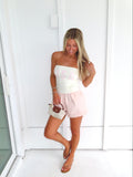 Woman wearing a strapless white top and pink shorts holding a purse against a white background.