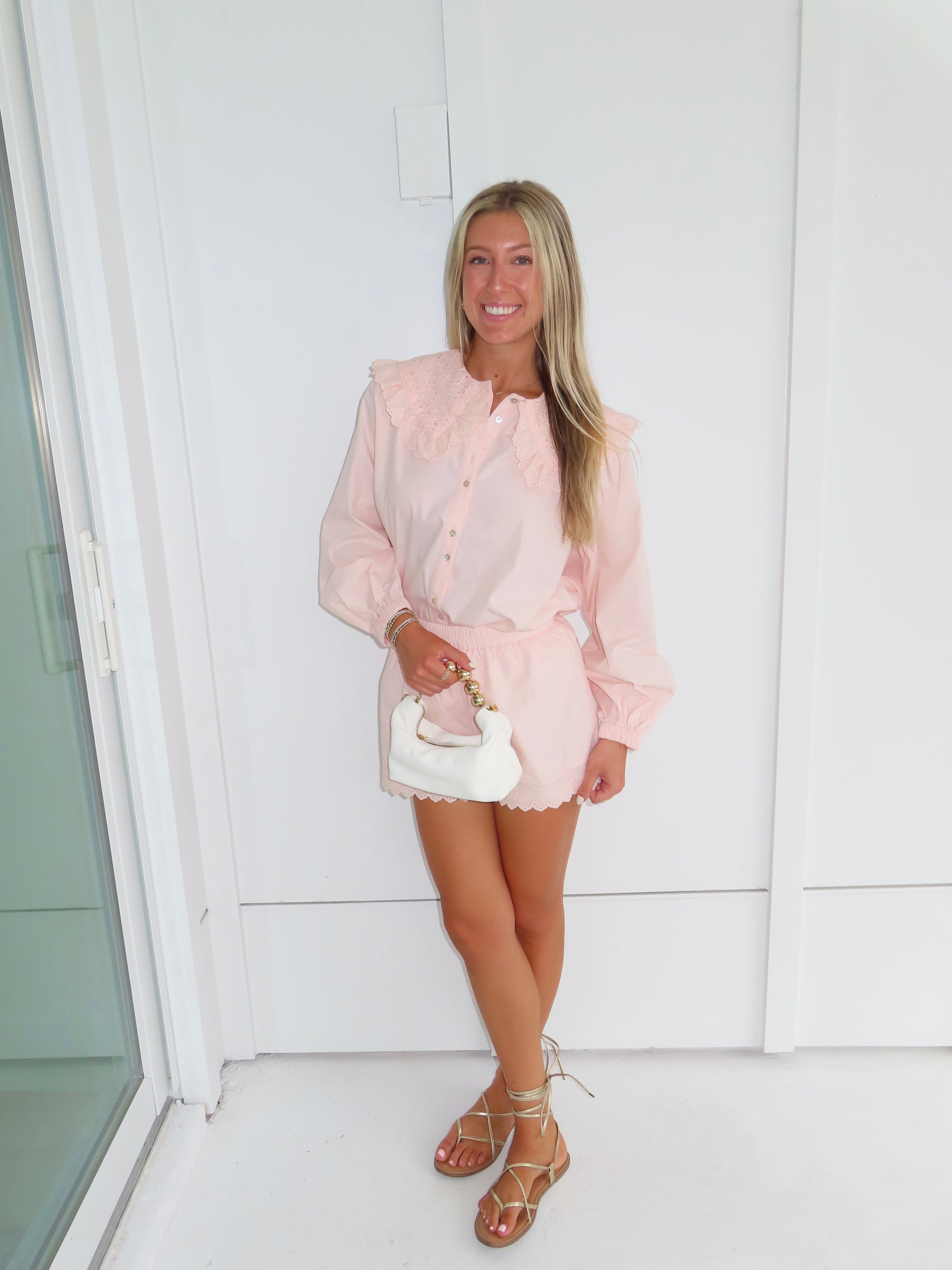 Woman in a light pink eyelet set holding a white handbag against a white wall.